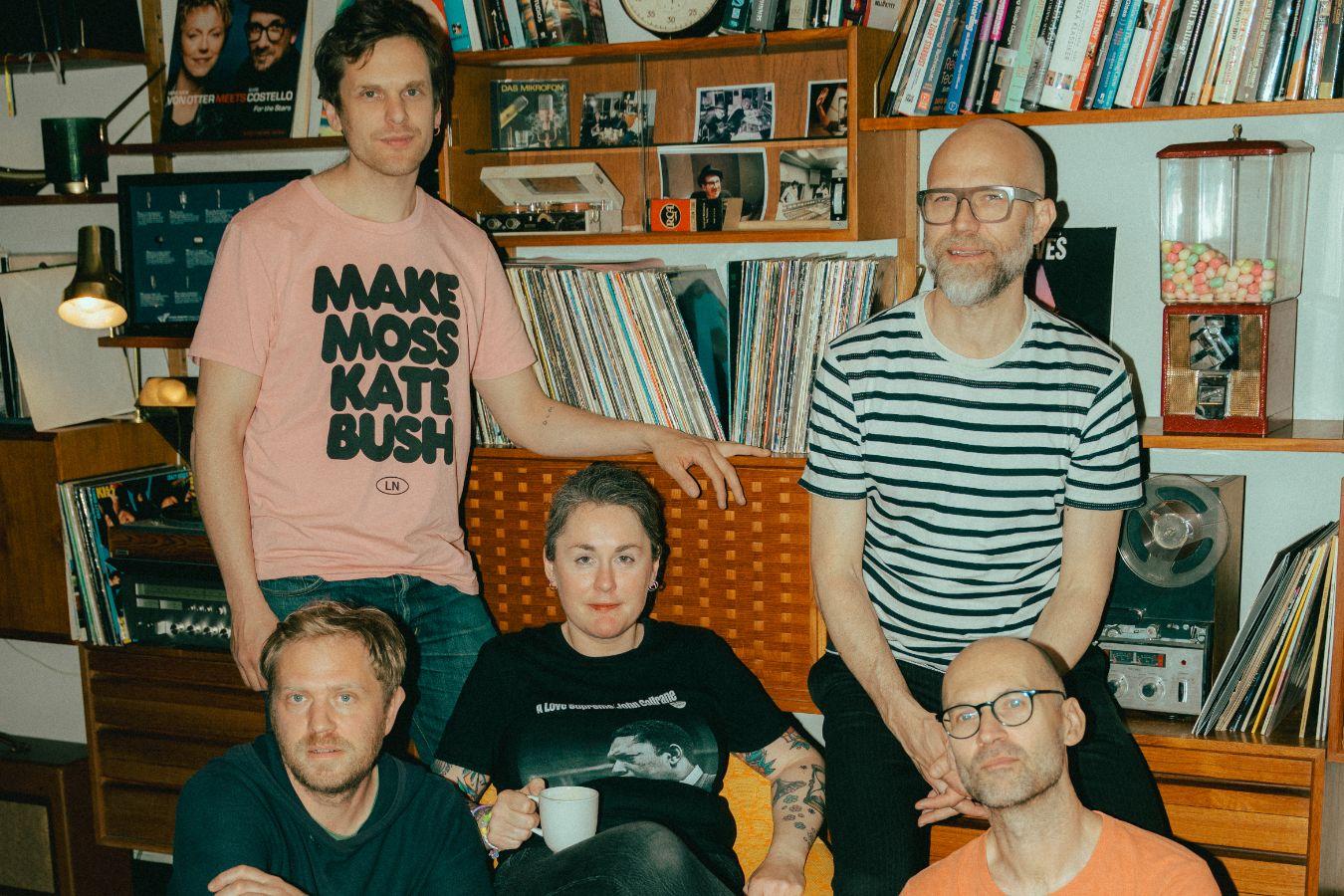 Gruppe von fünf Personen in einem Wohnzimmer mit Bücherregalen, Schallplatten und einem auffälligen T-Shirt in Rosa.