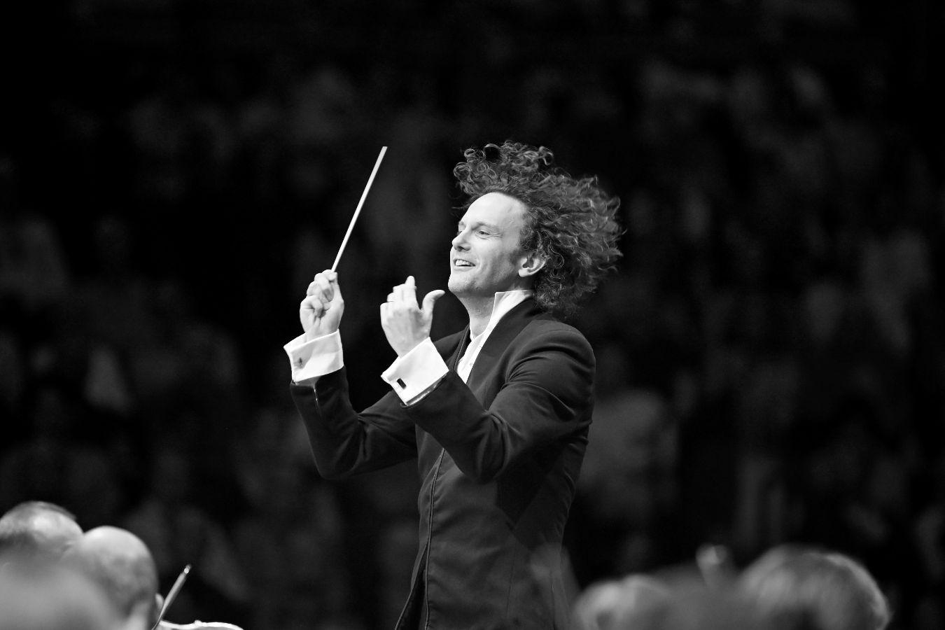 Ein Mann mit locken Haaren und einem Taktstock in der Hand dirigiert so überschwänglich, dass seine Haare herumwirbeln.
