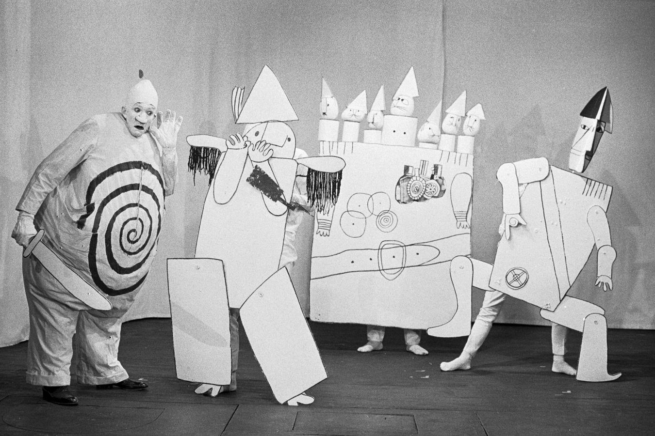 On a stage are people in costumes covered with painted paper, like in a comic strip.
