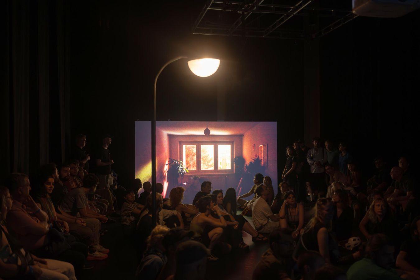 A dark room where people are sitting on the floor in front of a cinema screen, with a glowing streetlamp in the center.