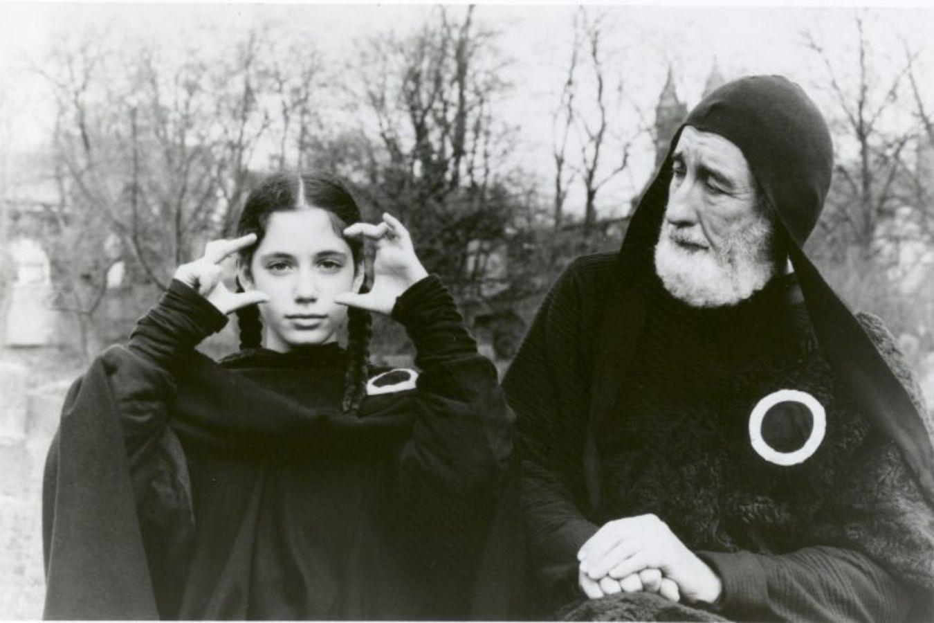 A girl forms a picture frame in front of her eyes with the thumbs and index fingers of both hands. An older man standing next to her looks at her. Both are wearing black cloaks.