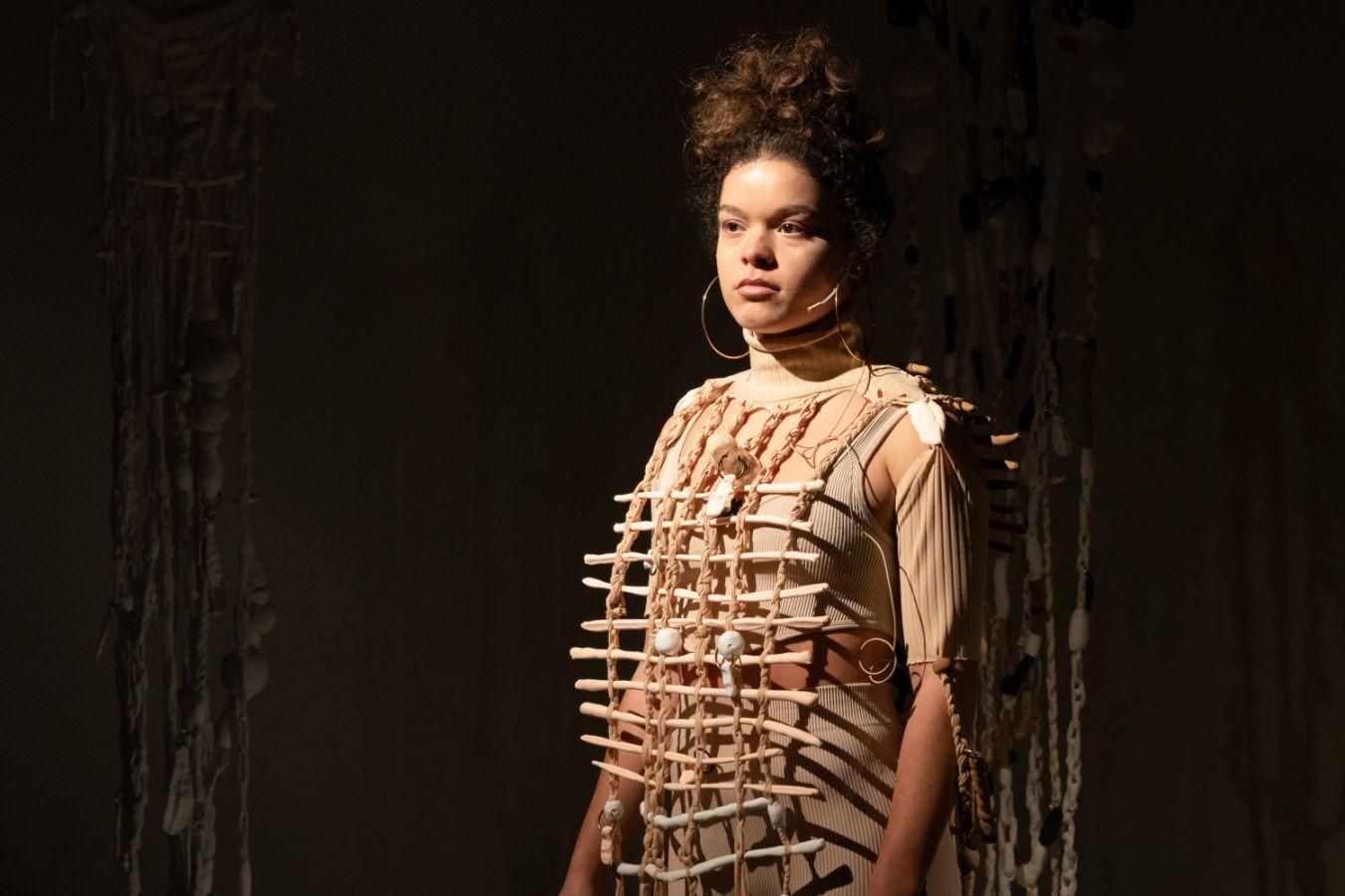 A photo of the artist Theresa Weber standing in a dark room with a spotlight on her. She is wearing an object on her chest that looks like it is made our of bones.