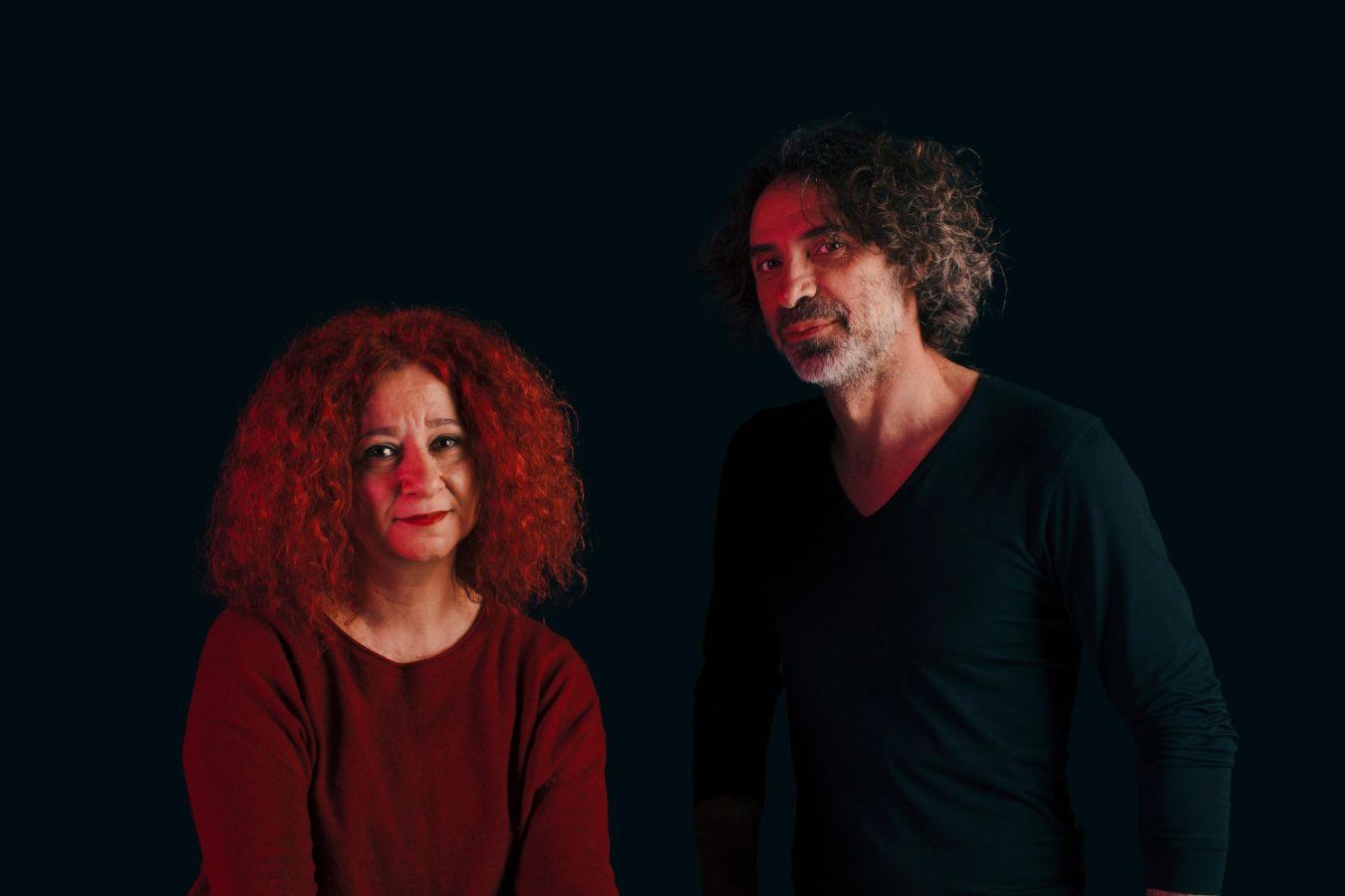 Zwei Personen sitzen bzw. stehen vor dunklem Hintergrund eine Person in rotem Oberteil, die andere in dunkler Kleidung.