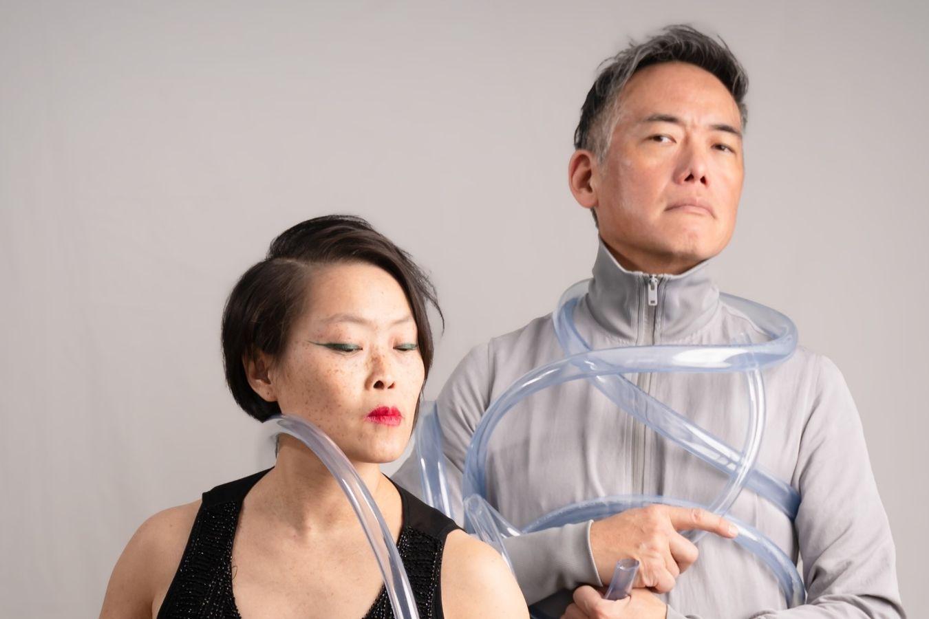 Eine Frau im schwarzen Kleid und ein Mann im grauen Anzug stehen vor einer grauen Wand. Ein langer durchsichtiger Schlauch ist teilweise um die beiden gewickelt.