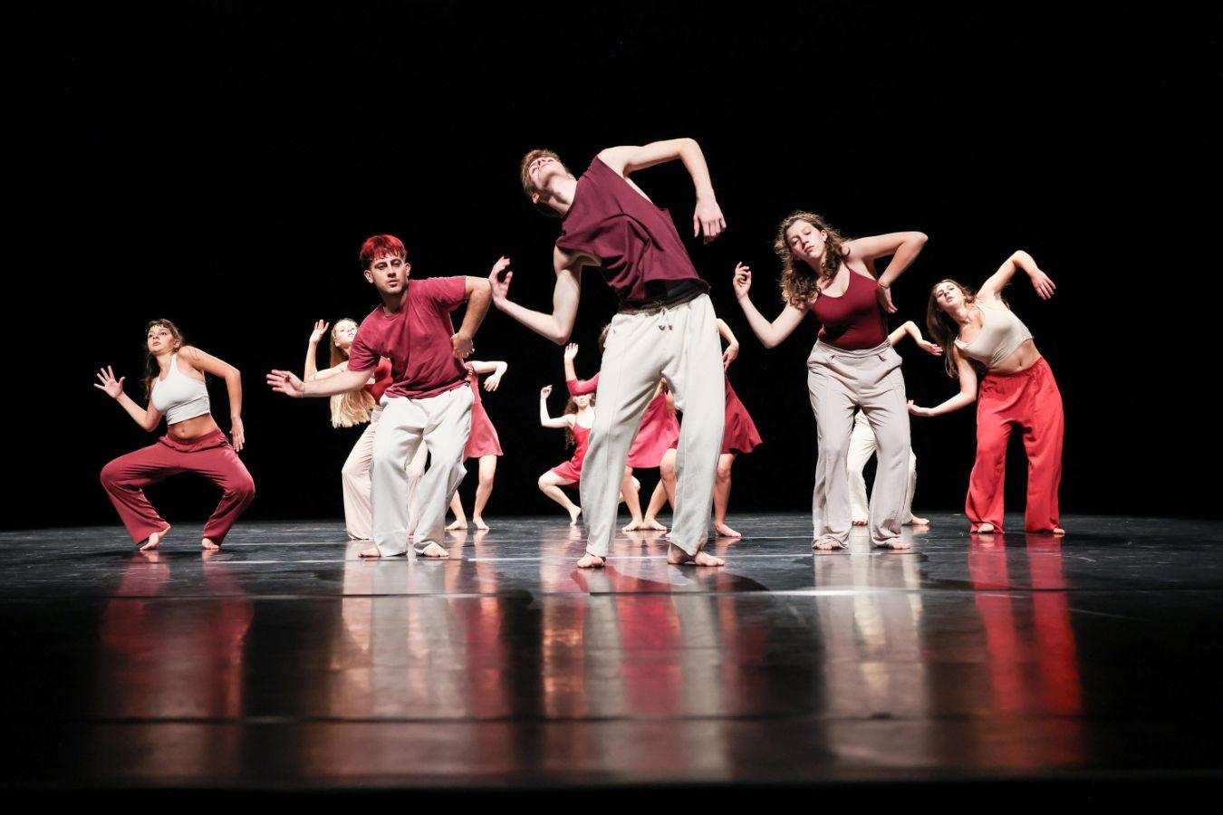 A group of dancers in red and light-coloured clothing performing a choreographed movement on a stage against a black background.