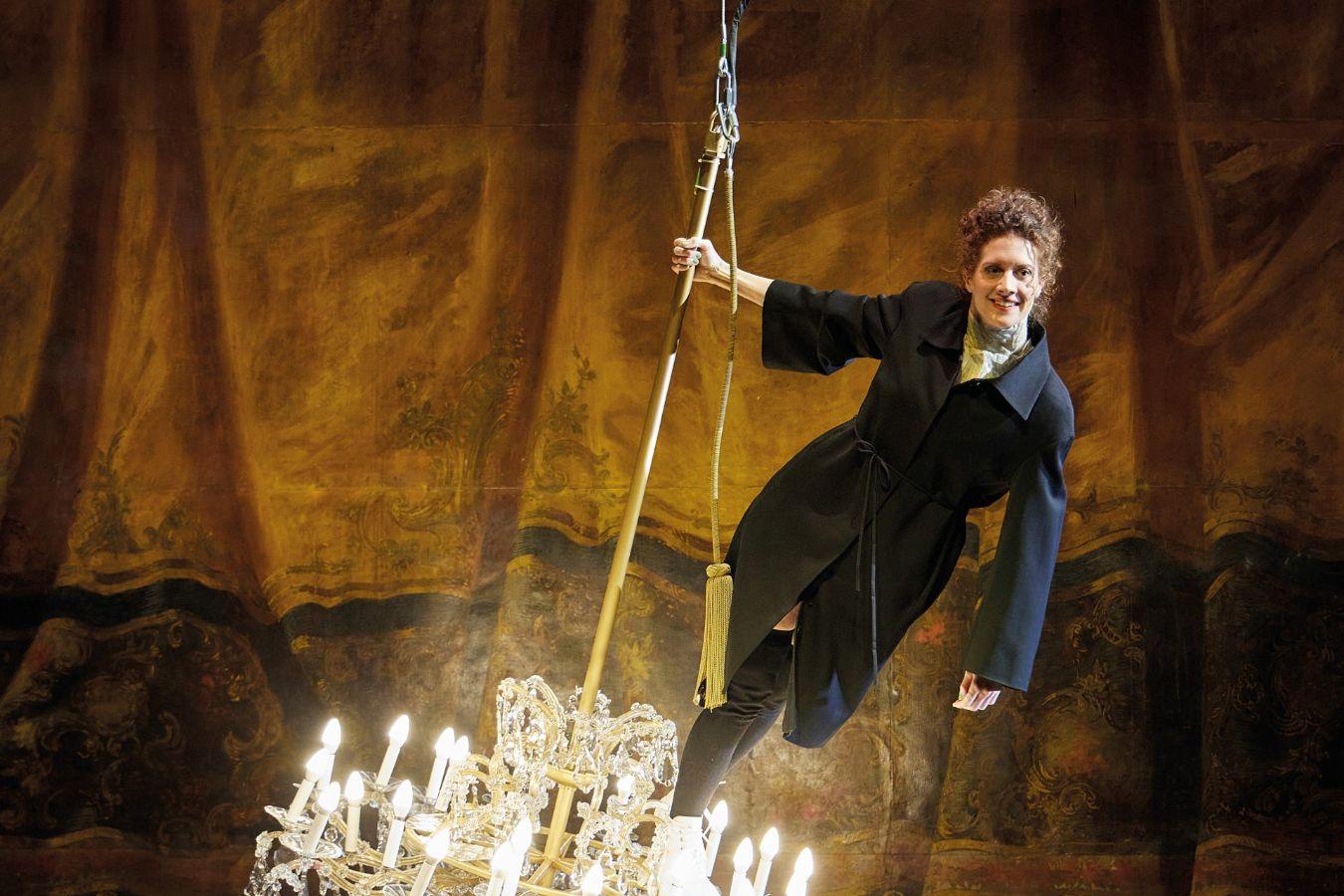 Eine Frau in einem Blazermantel steht lächelnd auf einem schwingenden Kronleuchter