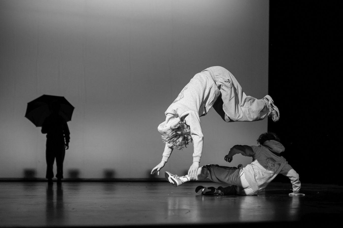 Schwarz-weißes Bühnenfoto: Im Fokus ein Tänzer in der Luft, Arme und Kopf im Abwärtsschwung. Im Hintergrund rechts sitzt eine Person mit dem rücken zur Kamera, links ist der Schatten einer Figur mit Regenschirm zu sehen.