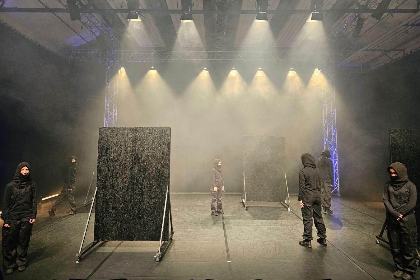 Tänzer*innen in schwarzer Kleidung stehen auf einer nebligen Bühne. Einige blicken nach vorne, andere sind von hinten zu sehen. Zudem befinden sich zwei schwarze Wandaufsteller auf der Bühne.