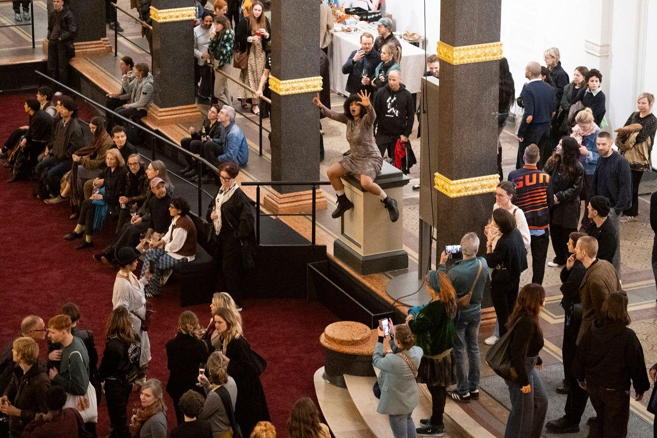 The full atrium of the Gropius Bau during the opening of the exhibition Ligia Lewis: I’M NOT HERE FORRRRR... The performer is seated on a pedestal in the center of the visitors.