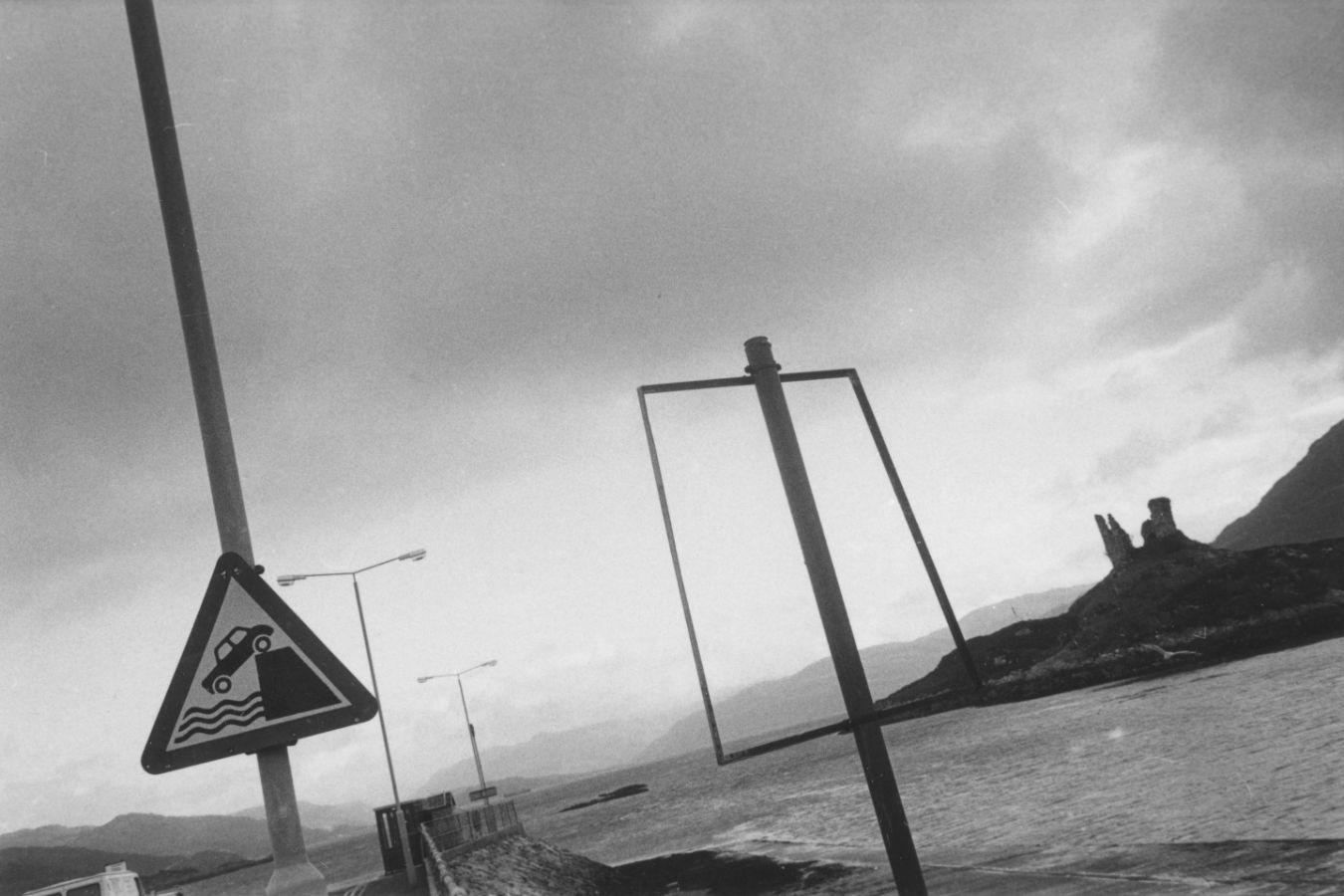 A warning sign depicting a coastline, the sea and a car falling into the sea stands on the sea wall of a bay. In the background are the sea and further headlands.
