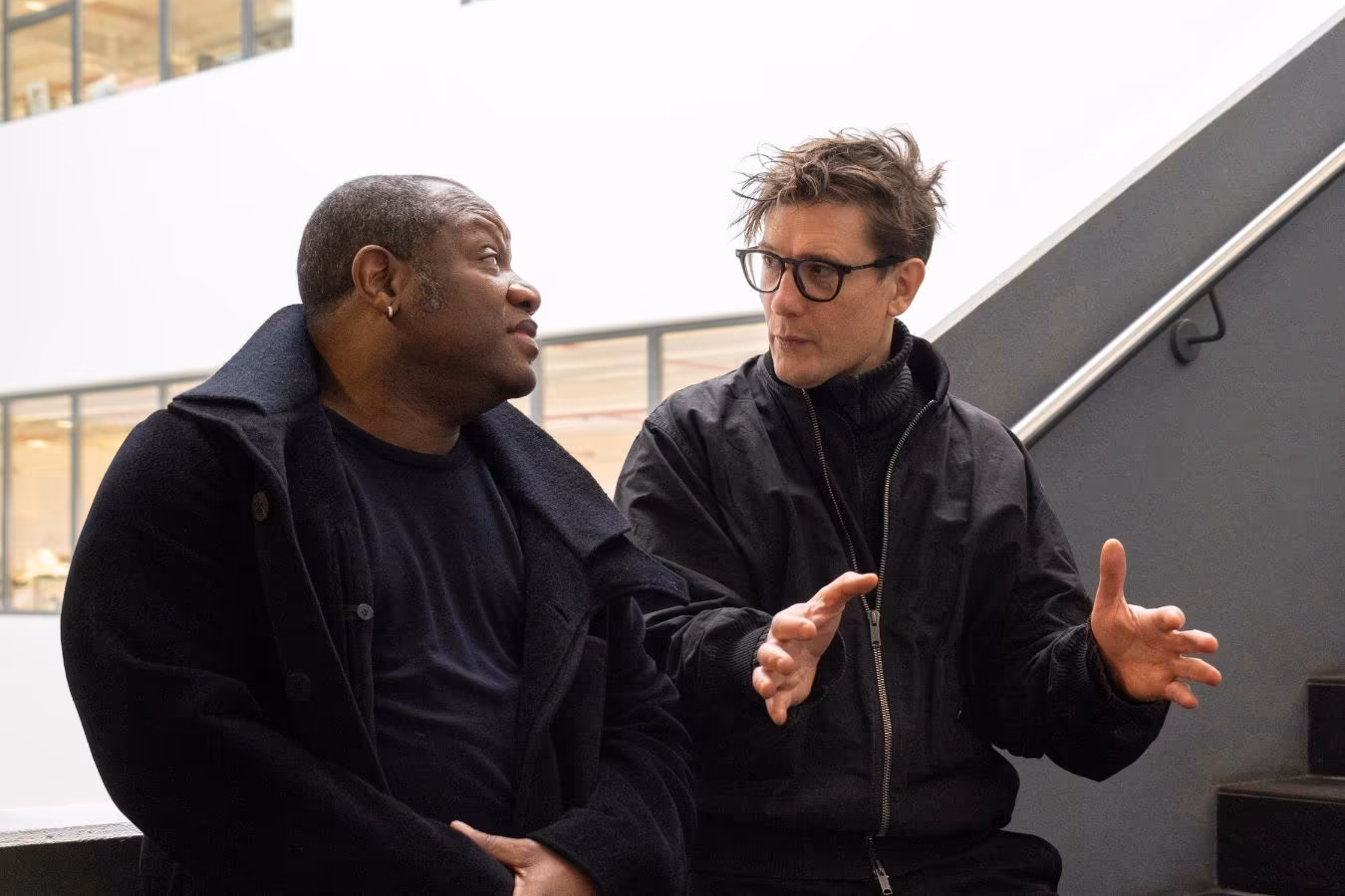 In einem Treppenhaus stehen die Künstler an ein Geländer gelehnt und sind ins Gespräch vertieft.