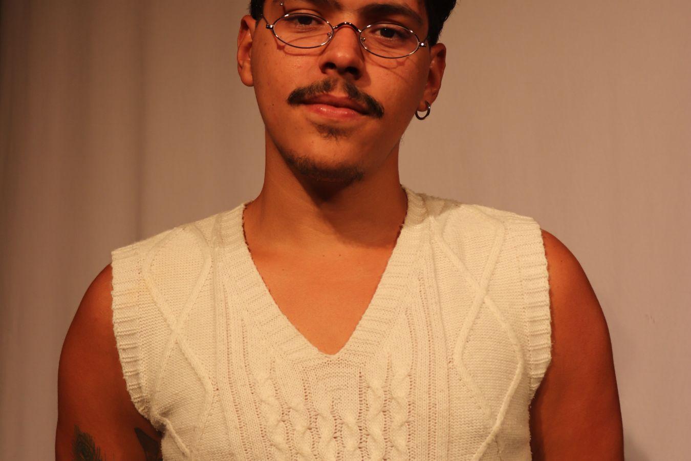A man with glasses and a beard wearing a white top standing with his arms crossed behind his back in front of a white wall