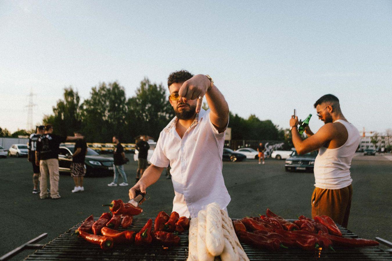 A shot of a person wearing a white shirt standing in front of a grill with red bell peppers and baguettes on it.