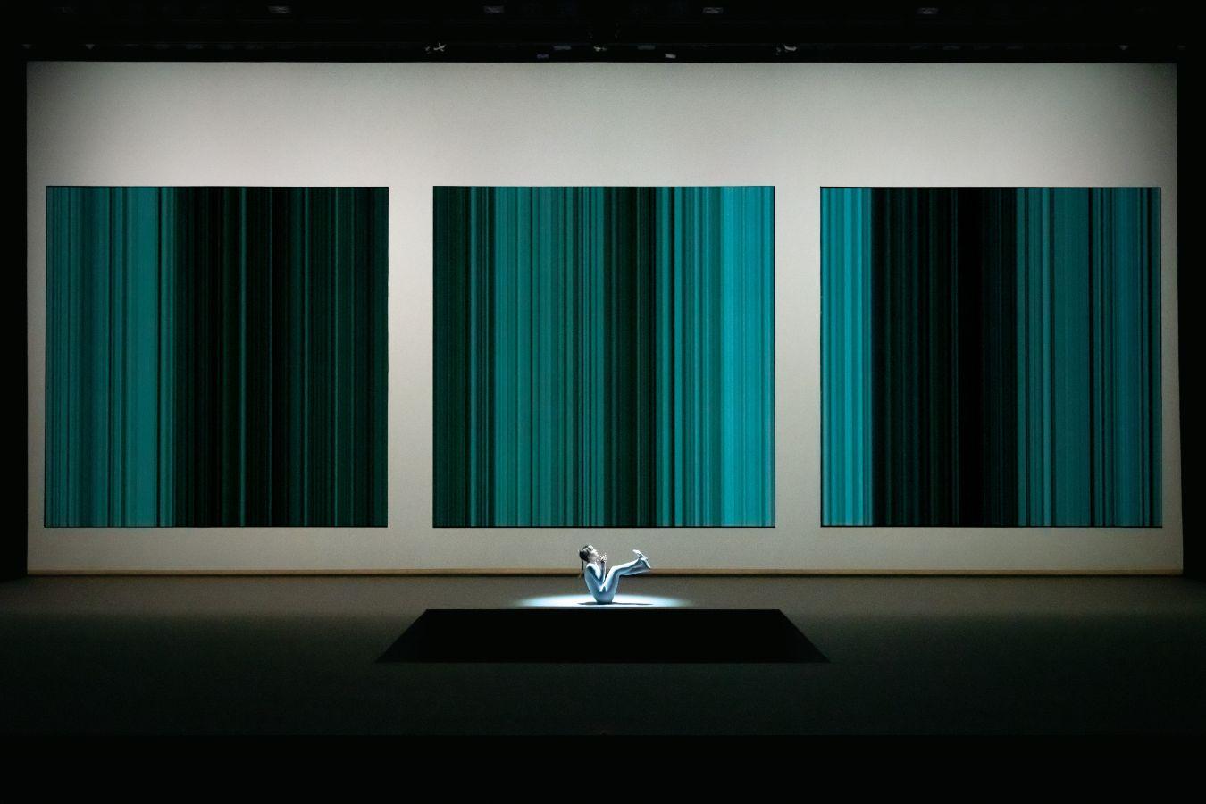 A person dressed in white sits behind a square opening in the stage floor; three screens are positioned behind them.
