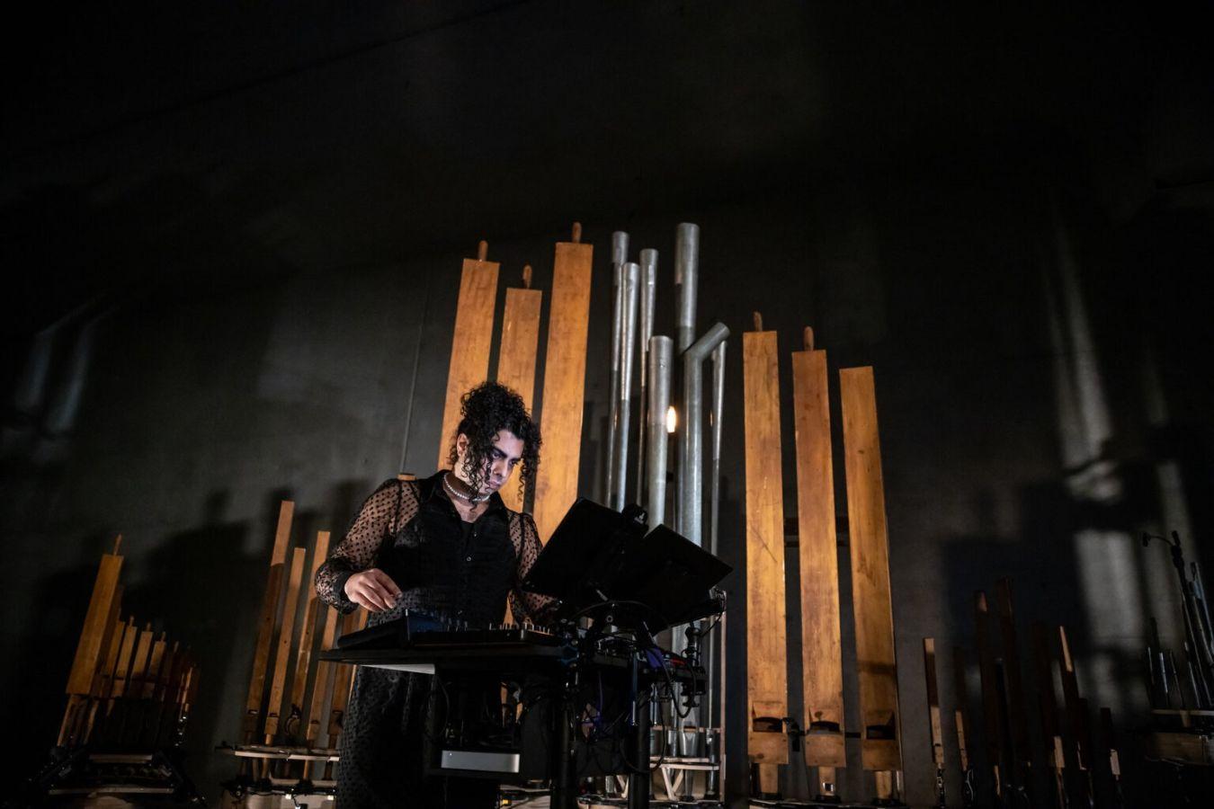 A person with high curly hair operates a mixing console. He stands in front of an installation made of organ pipes.