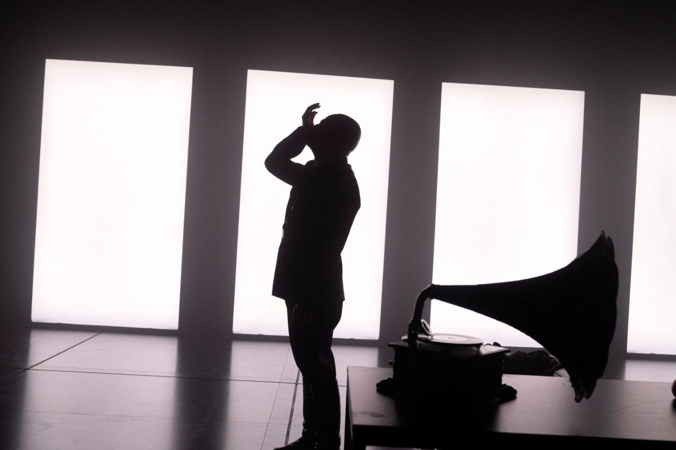 A man and a gramophone form silhouettes against brightly lit rectangular stage elements.