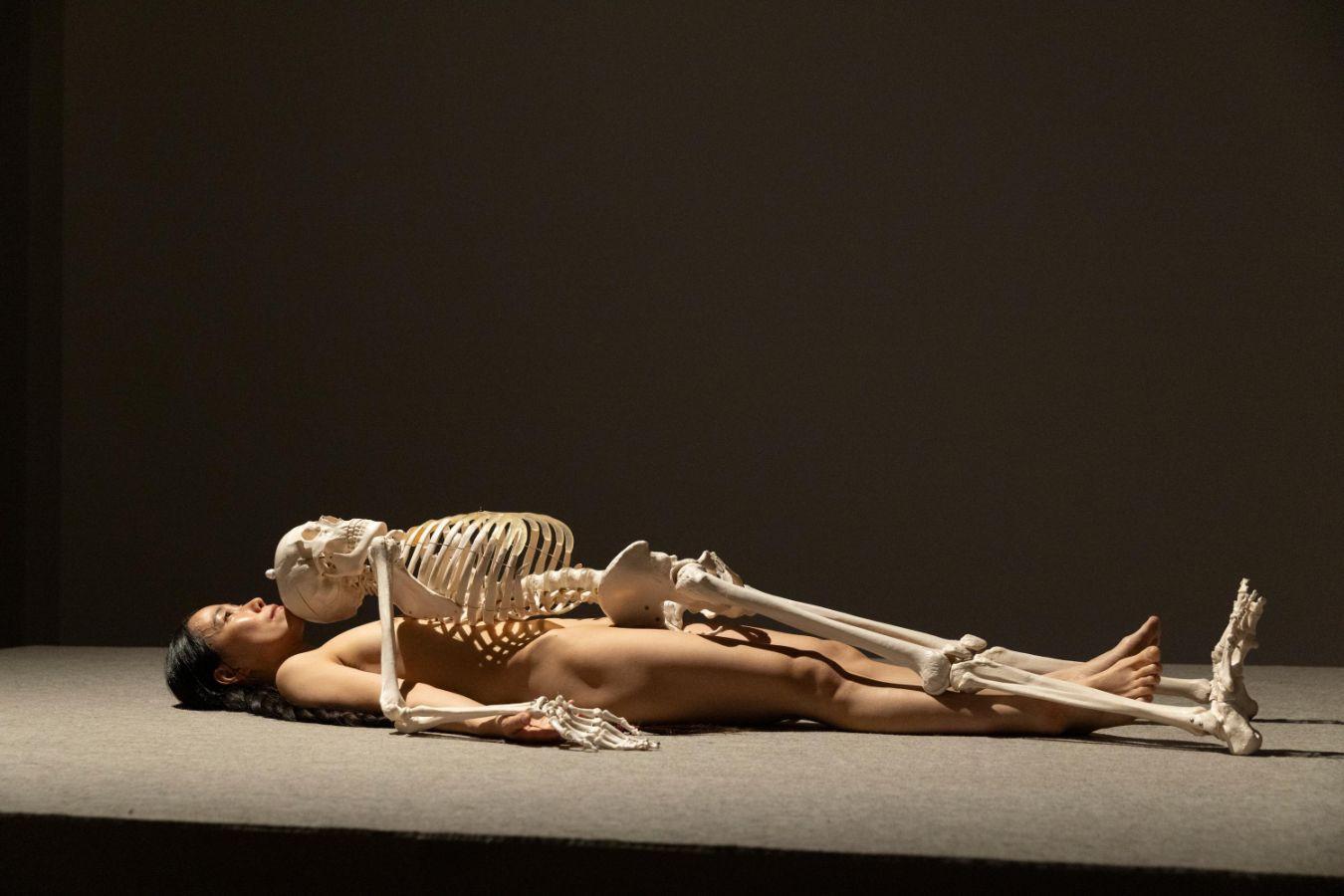 A naked person is lying on their back on an elevated surface. A skeleton is lying on top of the person.
