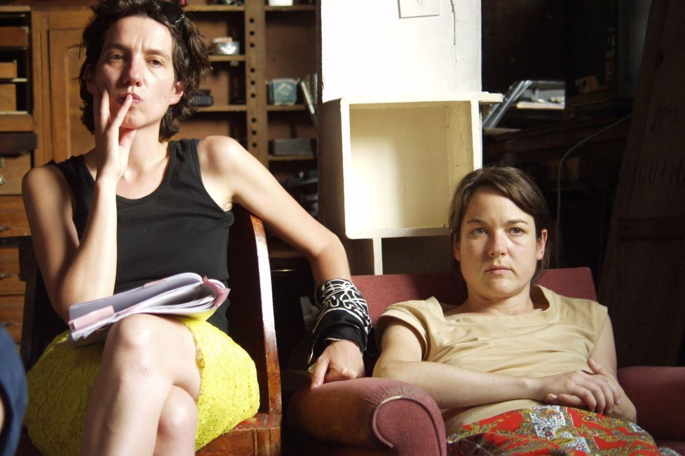 Zwei weiblich gelesene Personen sitzen auf zwei Sesseln, hinter ihnen ist ein Regal mit Büchern.