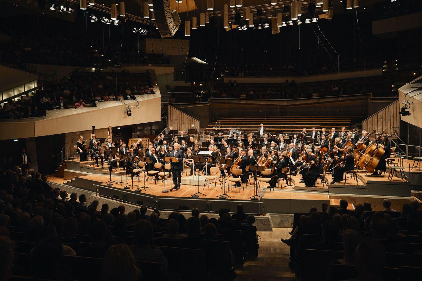 Großes Orchester auf einer hell ausgeleuchteten Bühne in einem Konzertsaal, mit Publikum im Vordergrund.
