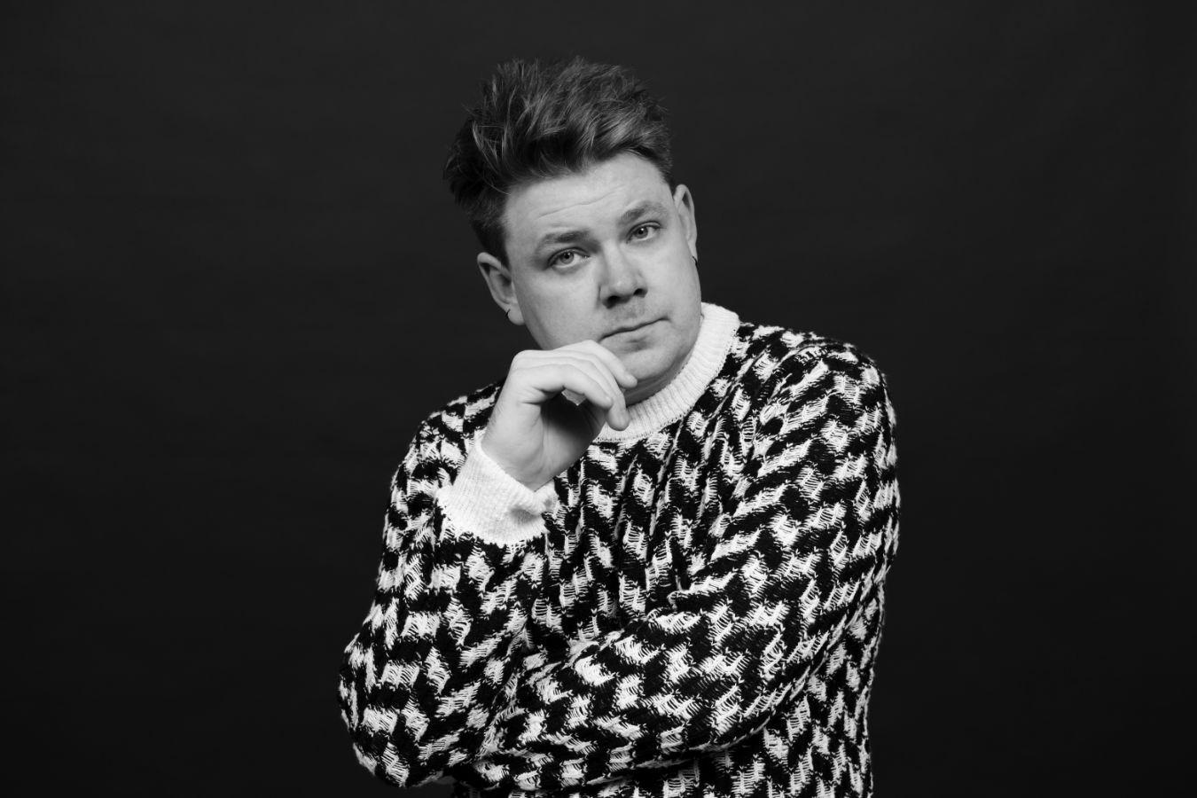 Simpson trägt einen Strickpulli und hält eine Hand ans Kinn.