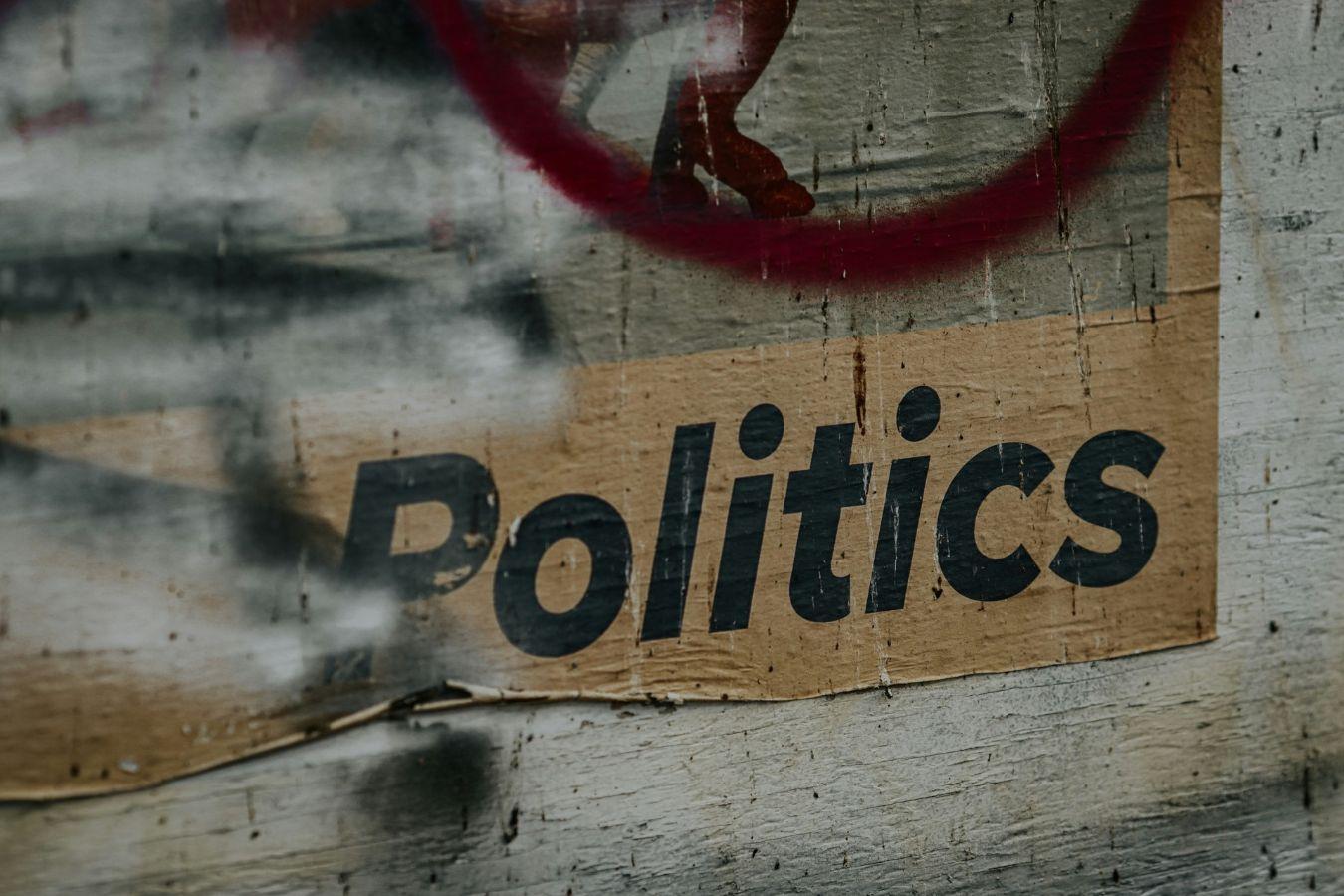 Close-up of a weathered surface showing the word “Politics” on cardboard, partially covered by paint.