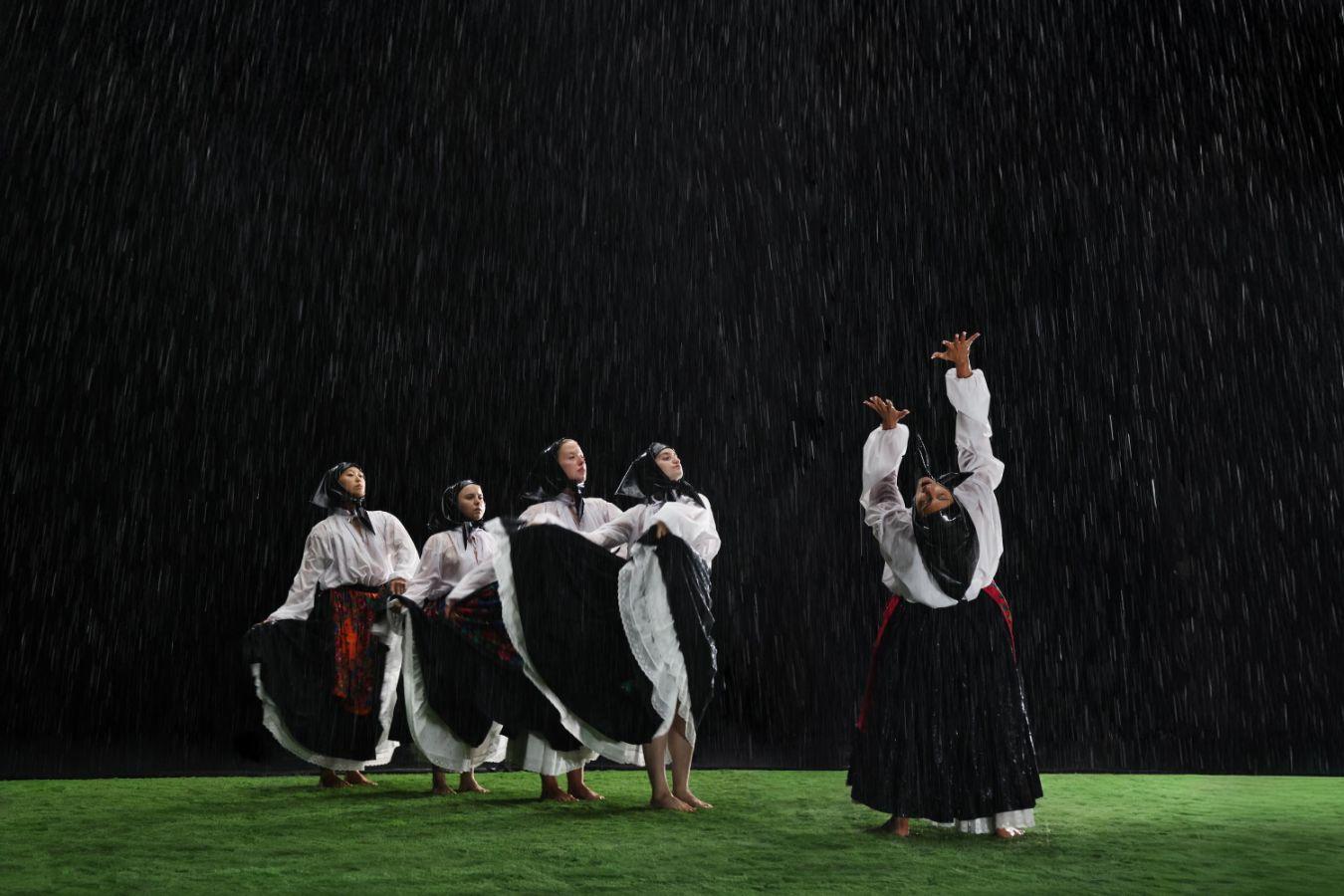 Fünf weiblich gelesene Personen mit schwarzem Kopftuch und knöchellangen Röcken bewegen sich auf einer grünen Wiese im Regen.