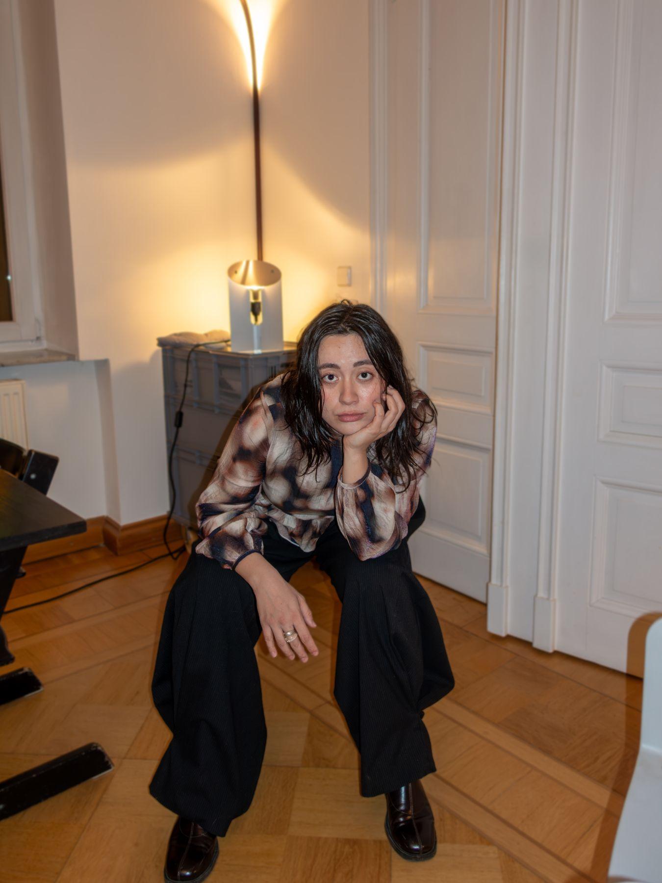 Eine Frau sitzt mit dunklen mittellangen Haaren und braun-gefleckter Bluse sitzt mit aufgestütztem Arm auf einem Stuhl