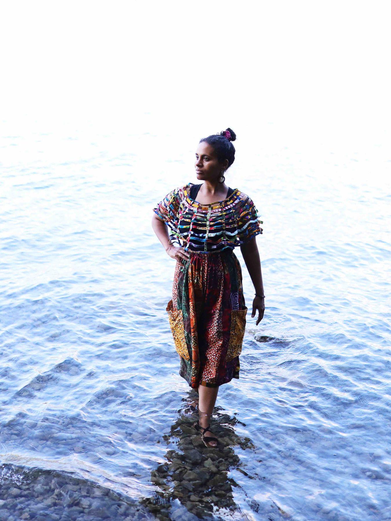 Eine Frau steht im Wasser und blickt in die Ferne. Sie trägt bunte Kleidung.