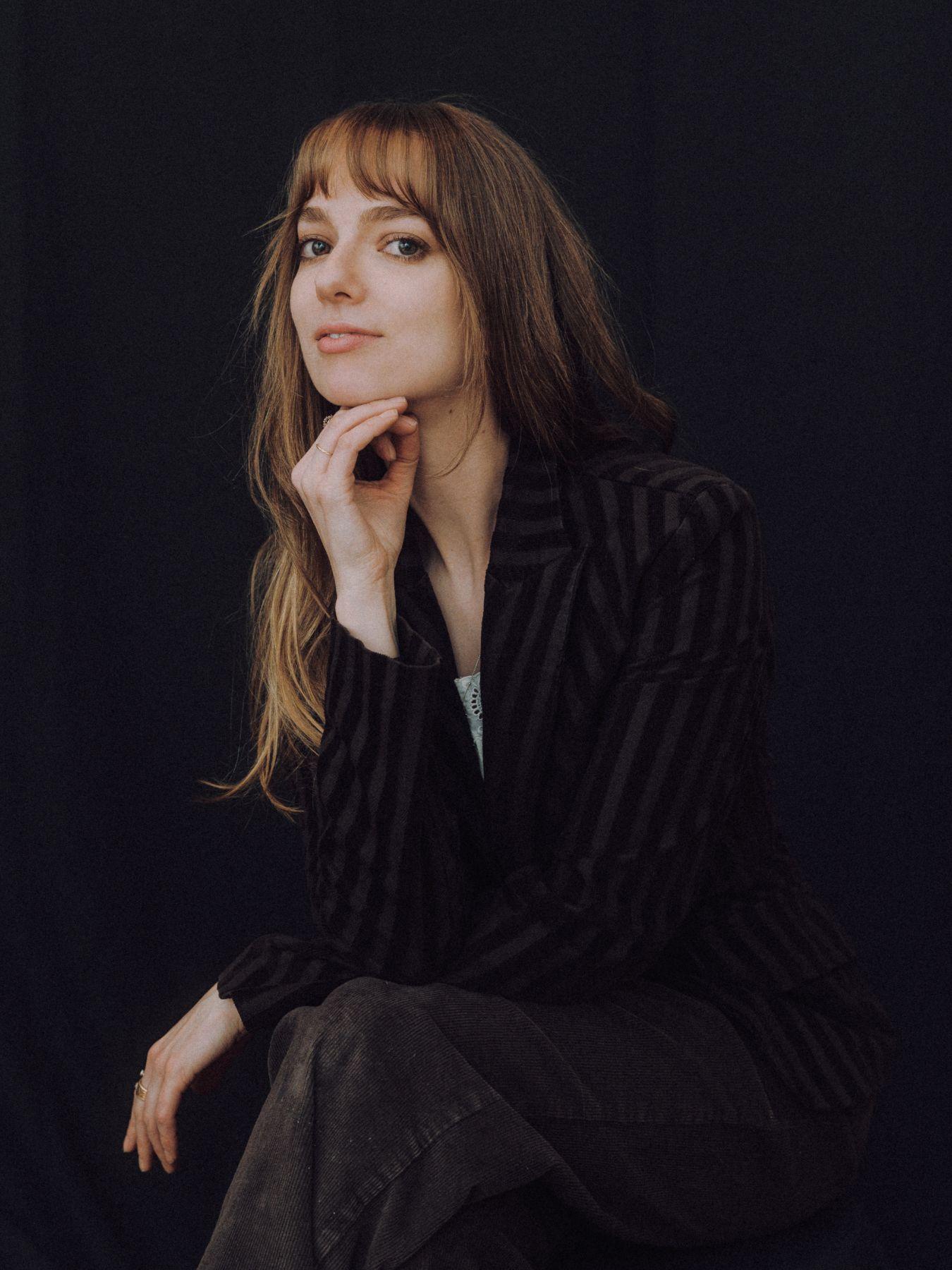 A woman with long light-brown hair in a black blazer sitting and resting her head on her hand
