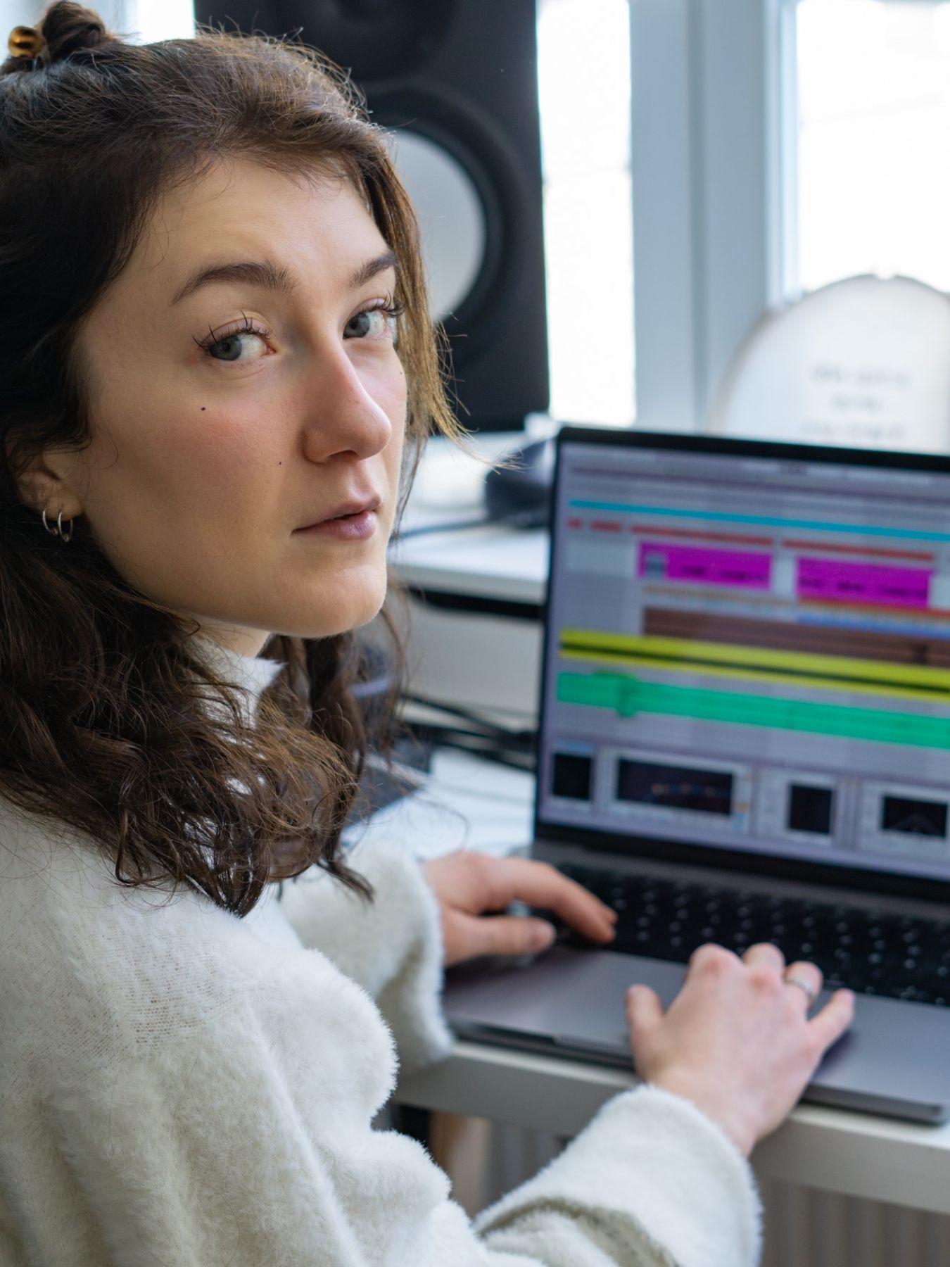 Eine junge Frau sitzt am Laptop und blickt über die Schulter direkt in die Kamera. Sie trägt einen hellen Pullover.