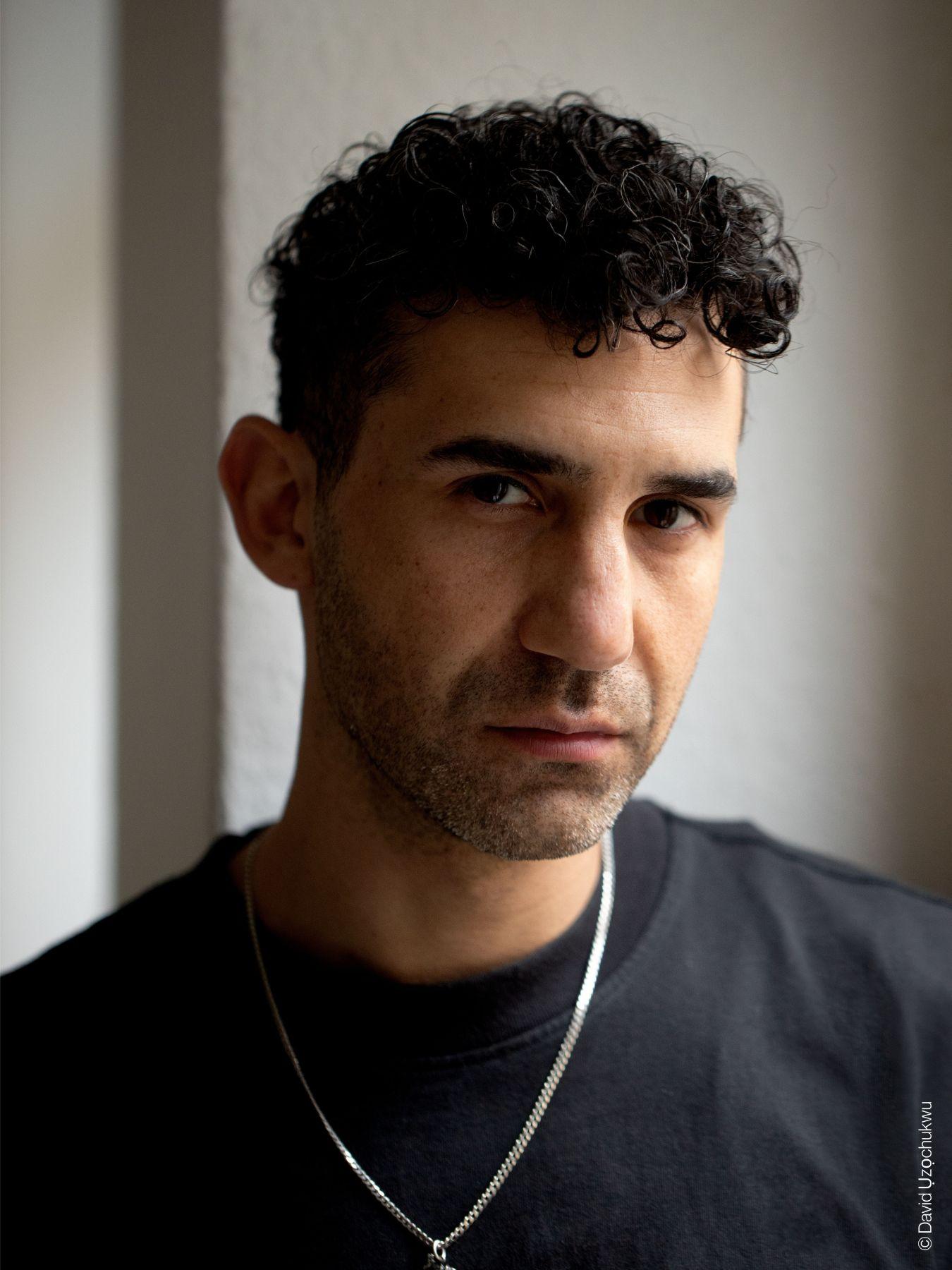 A young man with dark curly hair, a black T-Shirt and a silver necklace