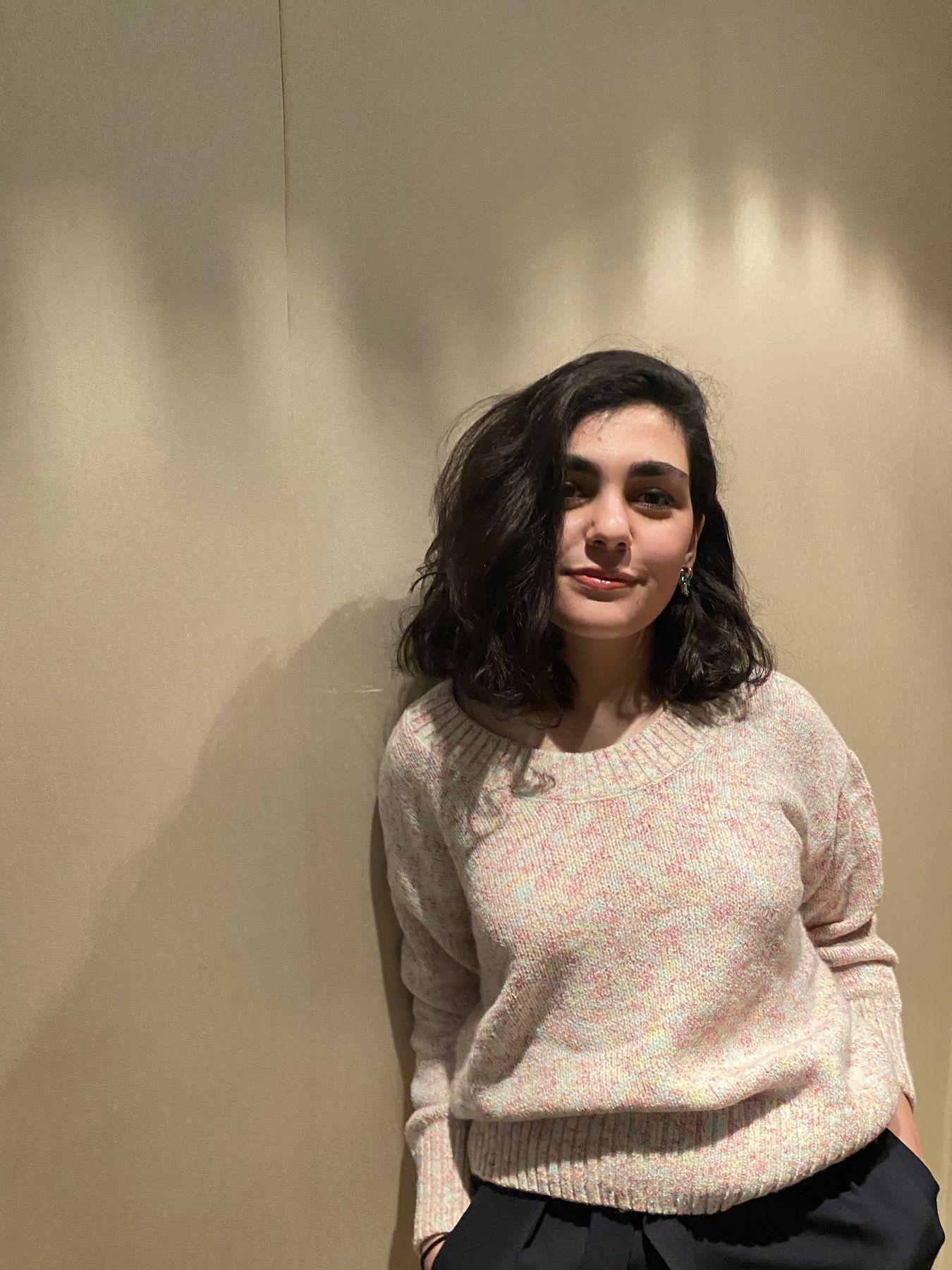 A woman with dark-brown hair and a beige jumper leaning against a beige wall