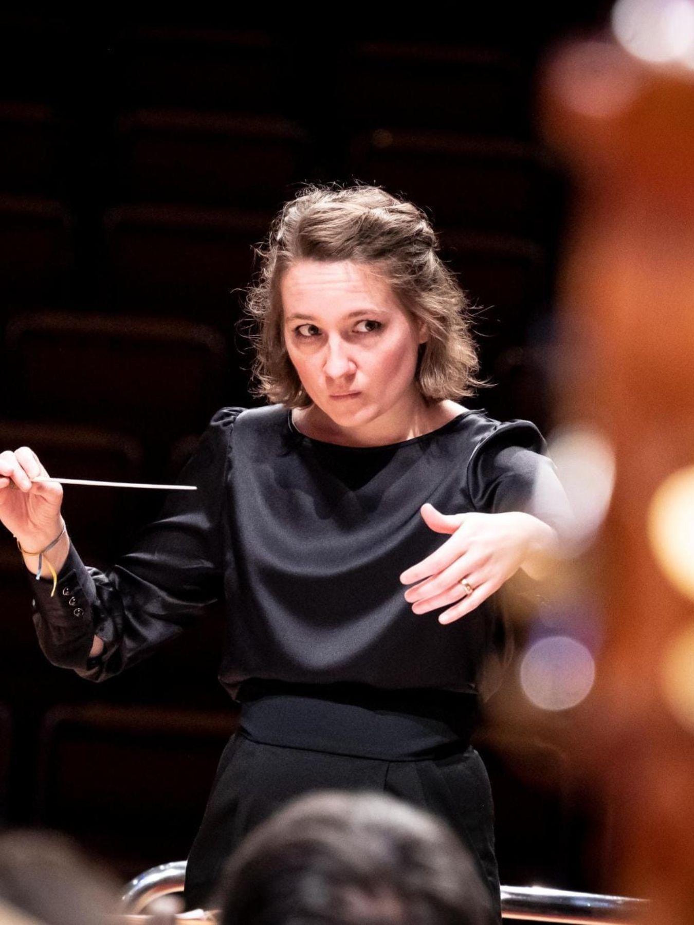 Eine Dirigentin steht vor dem Orchester und gibt mit dem Taktstock in der linken Hand den Einsatz.