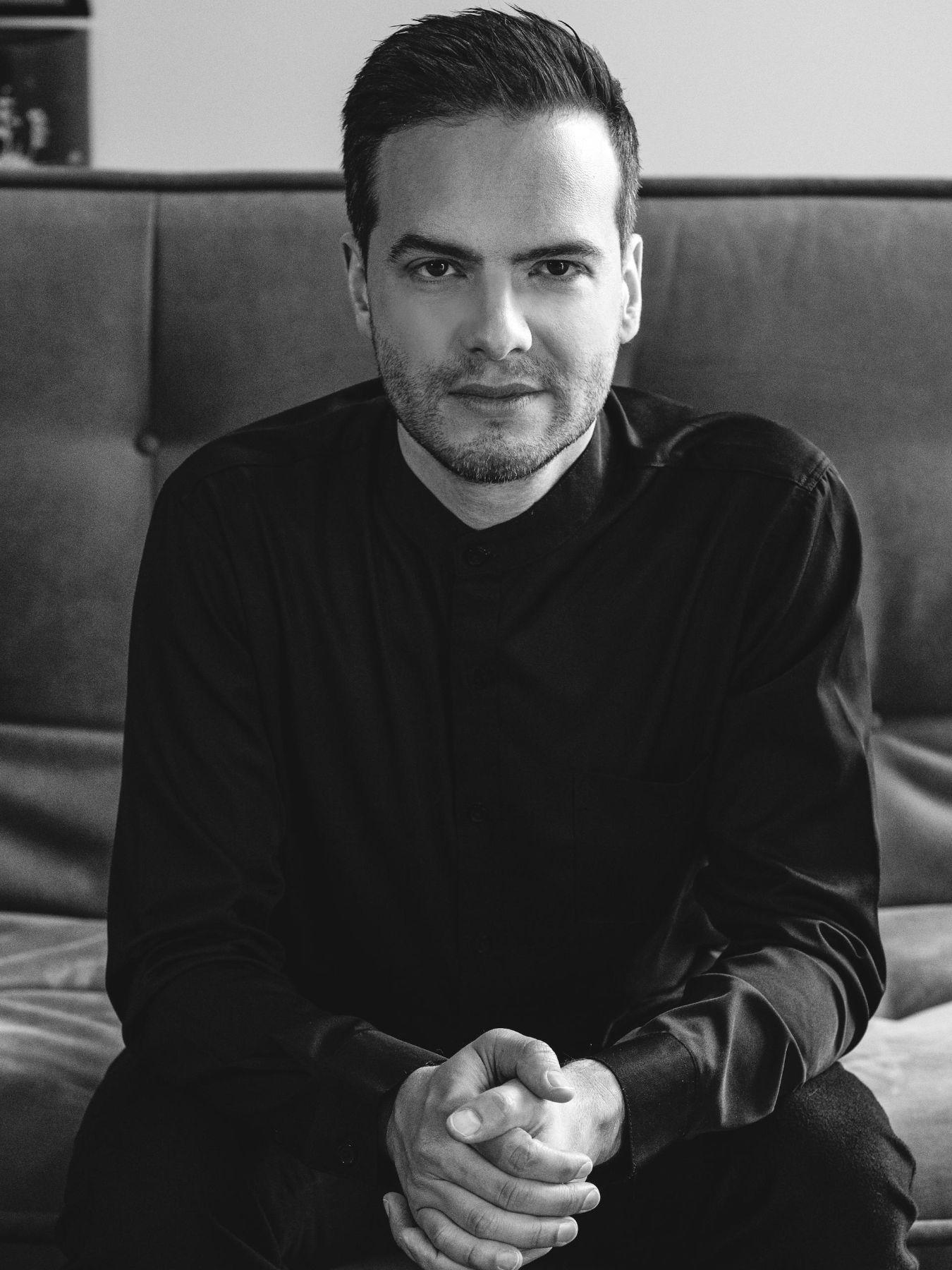 A man is sitting on a sofa with his arms crossed, looking at the camera.