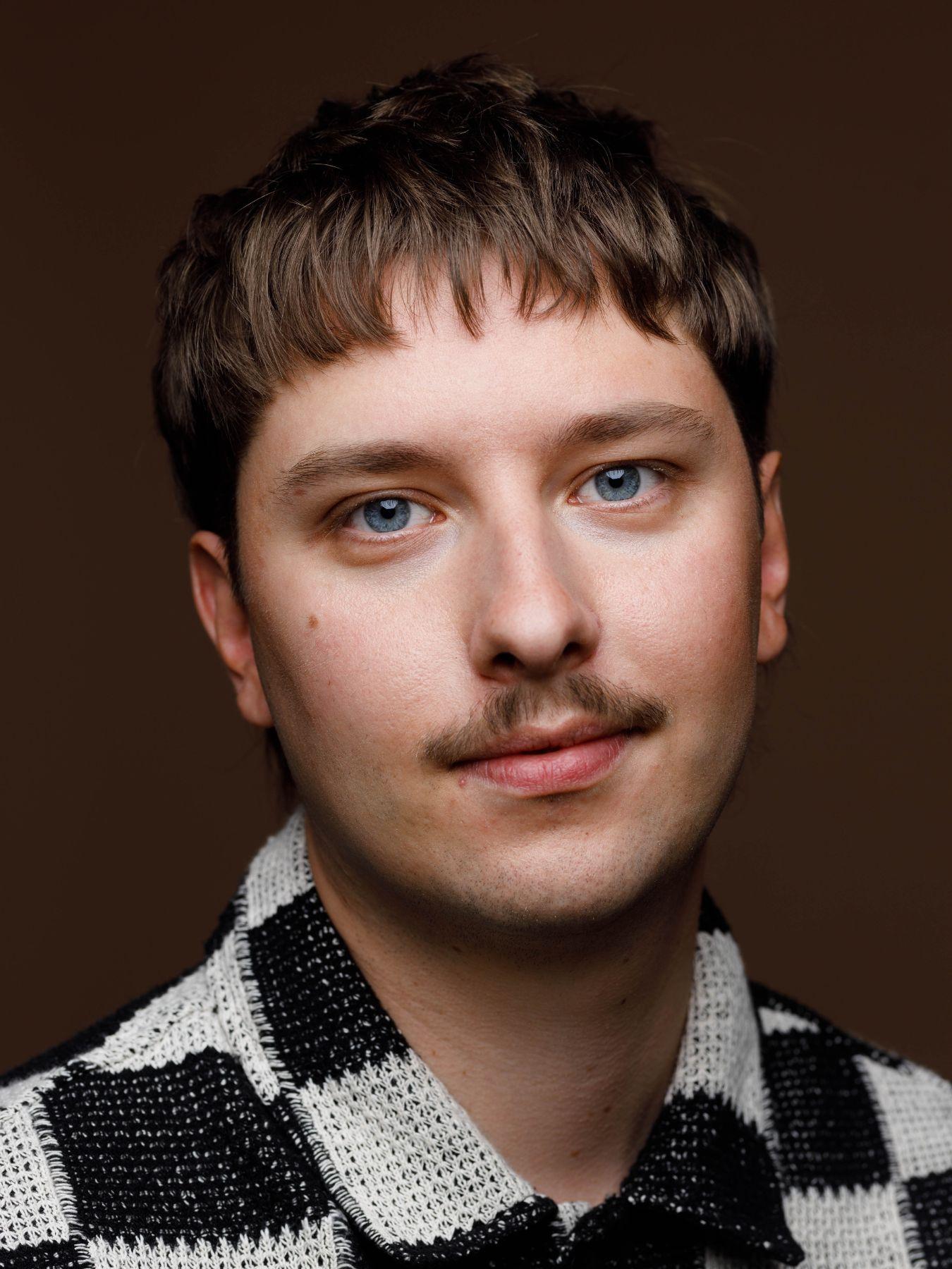 A man with a moustache and a checked shirt in front of a neutral background