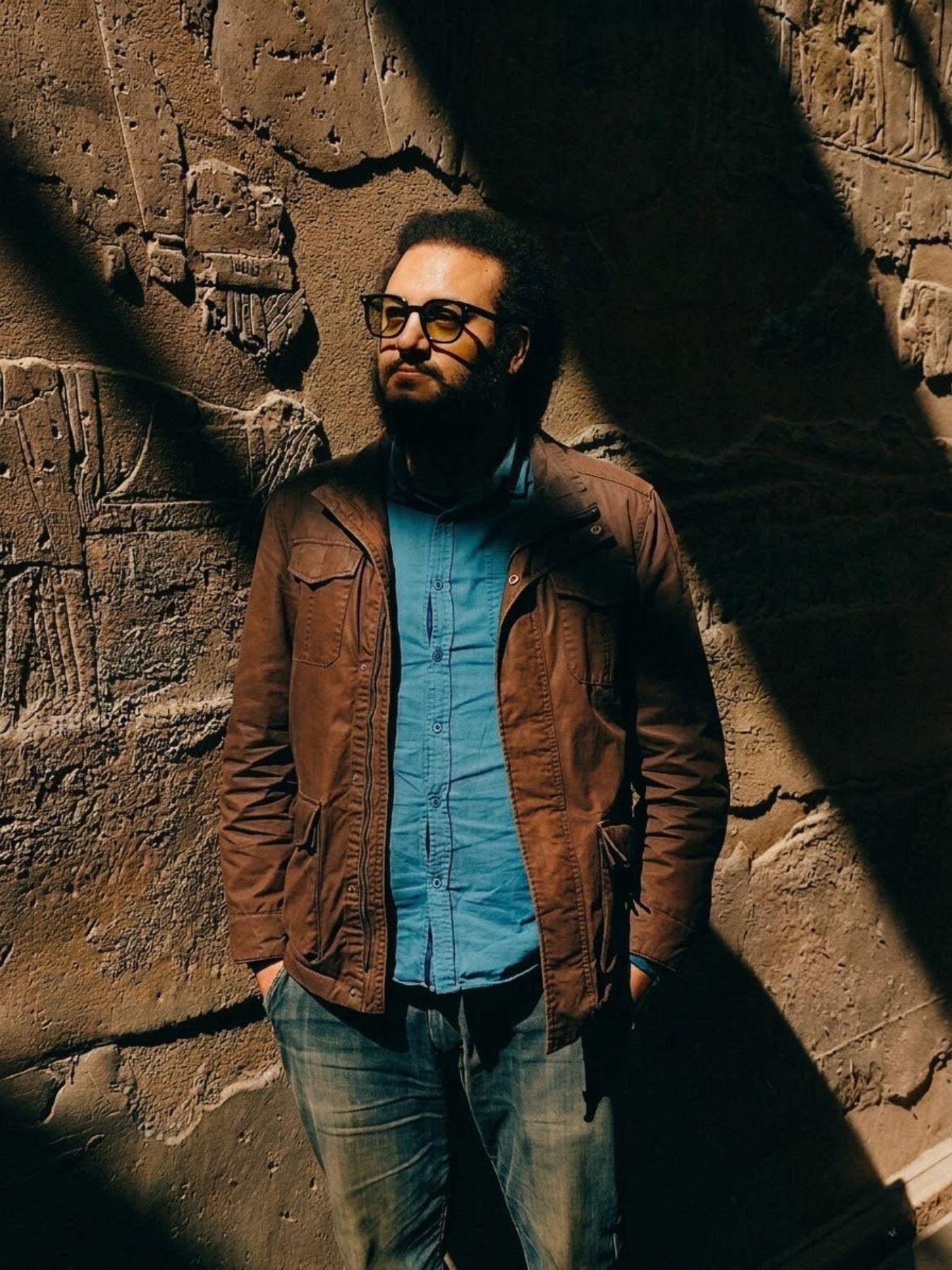 A man with dark brown hair, a beard and glasses leaning against a stone wall with his hands in his pockets