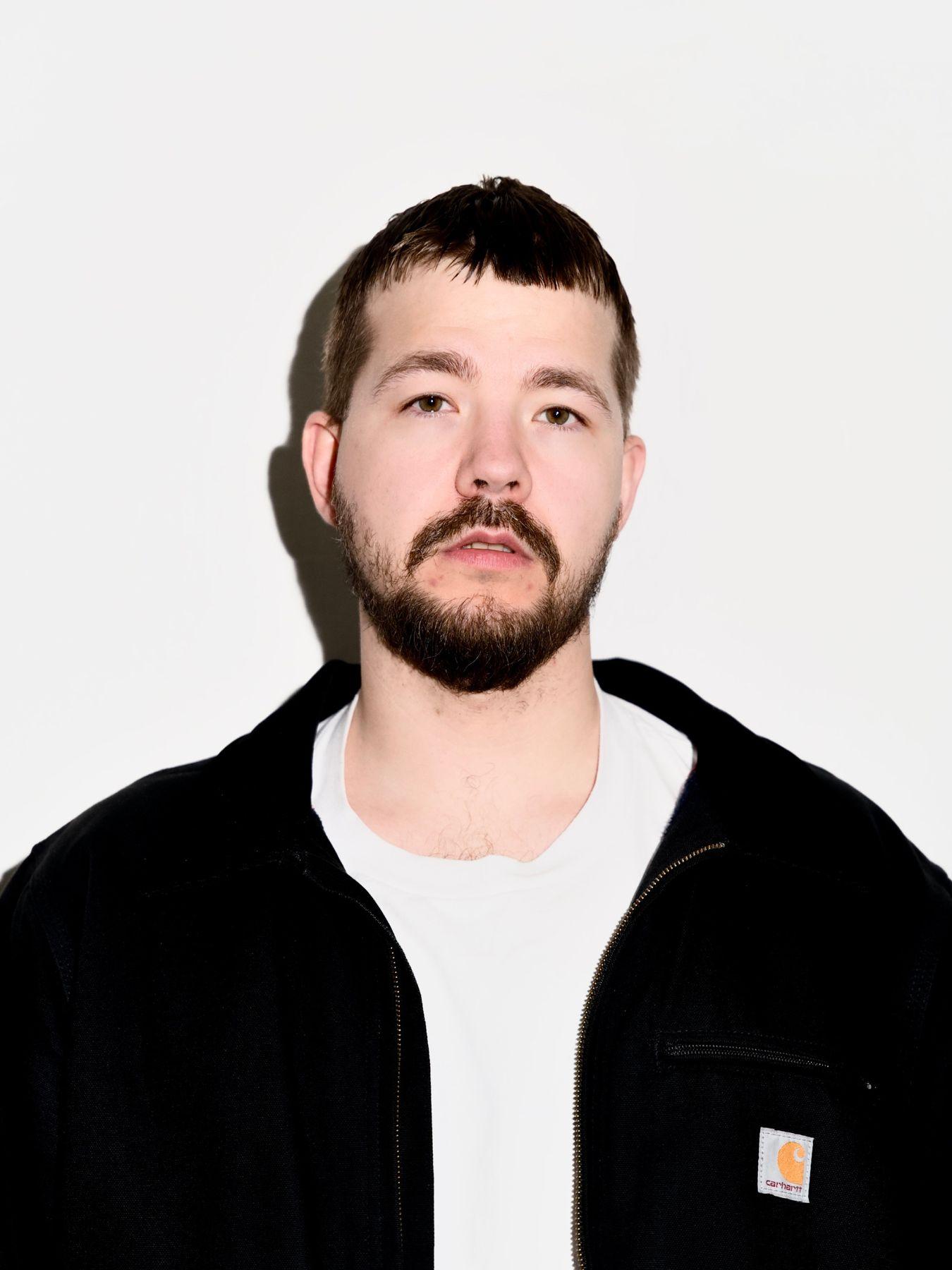 A man with short brown hair and a beard wearing a white top and a black jacket