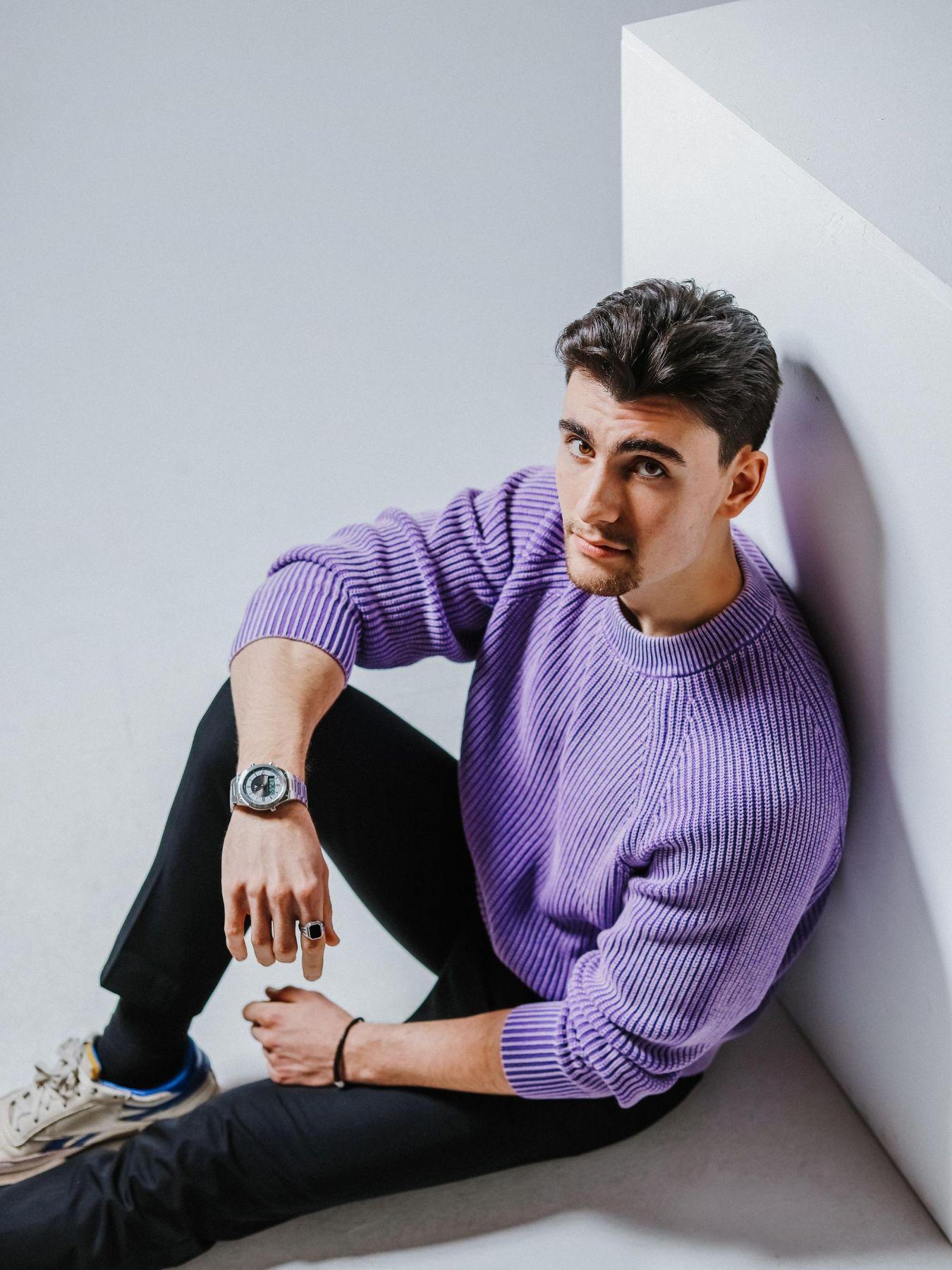 Eine Person mit kurzem dunkelbraunen Haar und einem lila Pullover sitzt gegen eine Wand gelehnt und schaut nach oben.