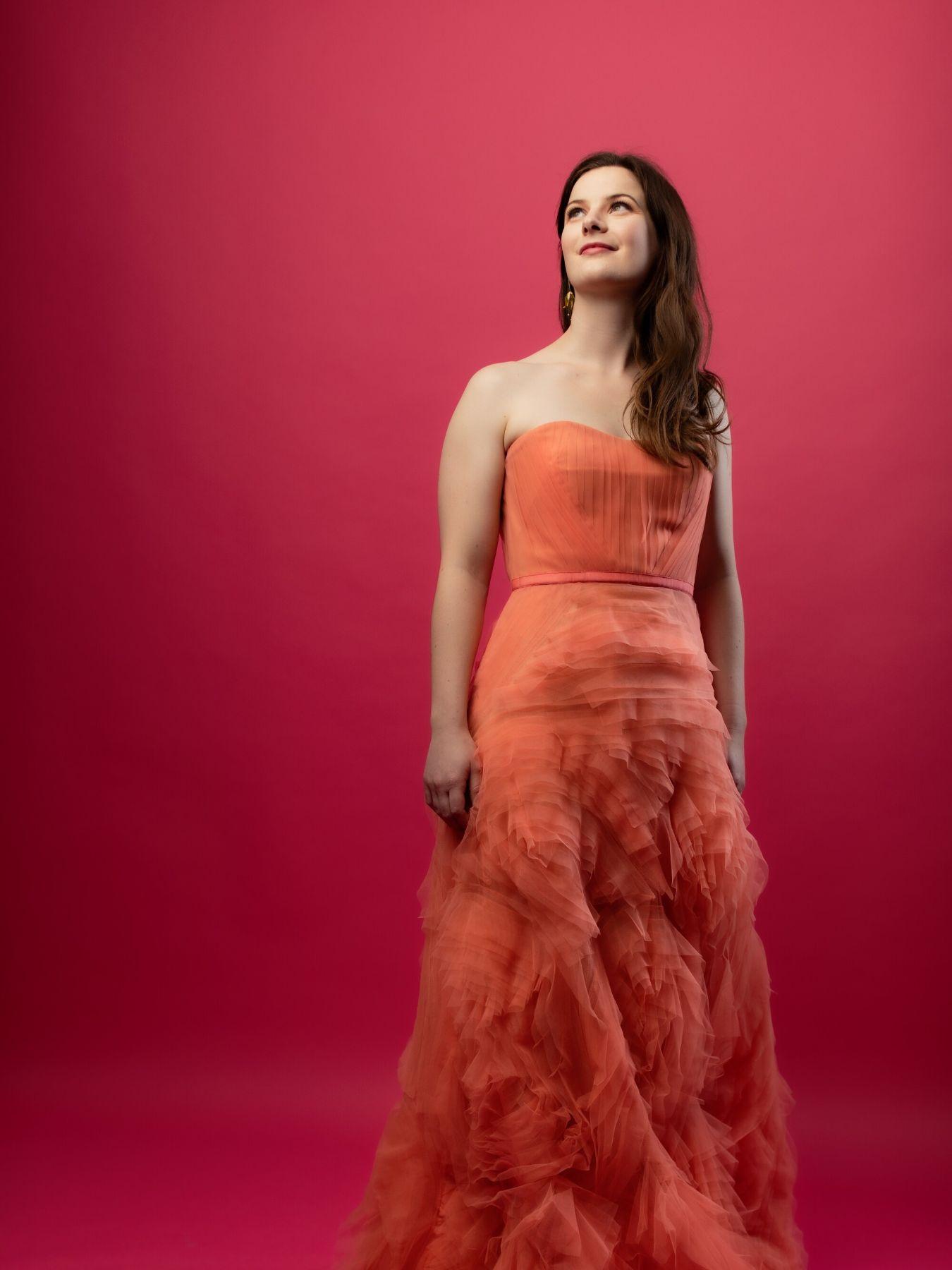 A woman with long brown hair stands in a long, elegant red dress in front of a dark red wall and looks upwards.