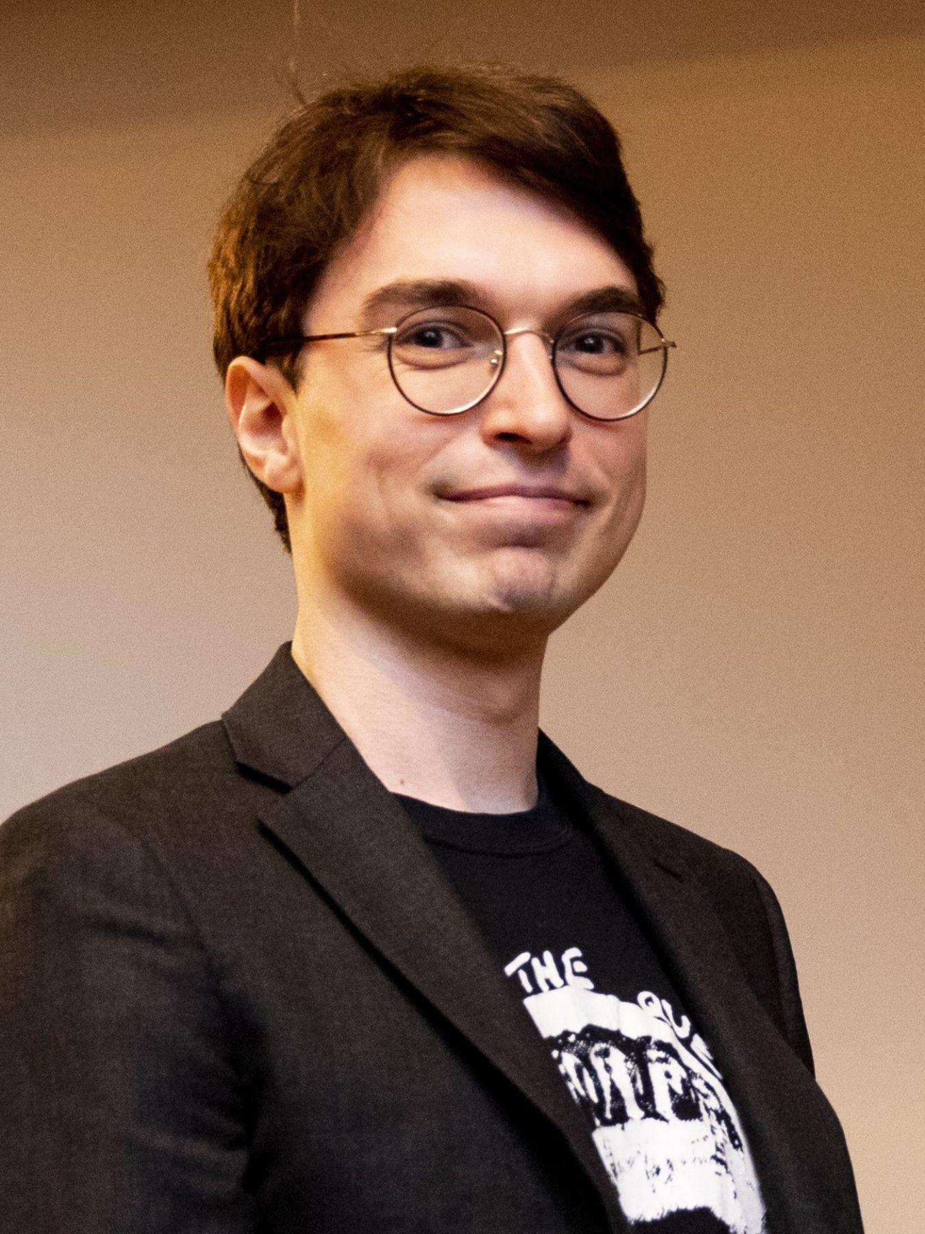 Eine Person mit kurzem braunen Haar und Brille. Die Person trägt ein Shirt und Sakko.