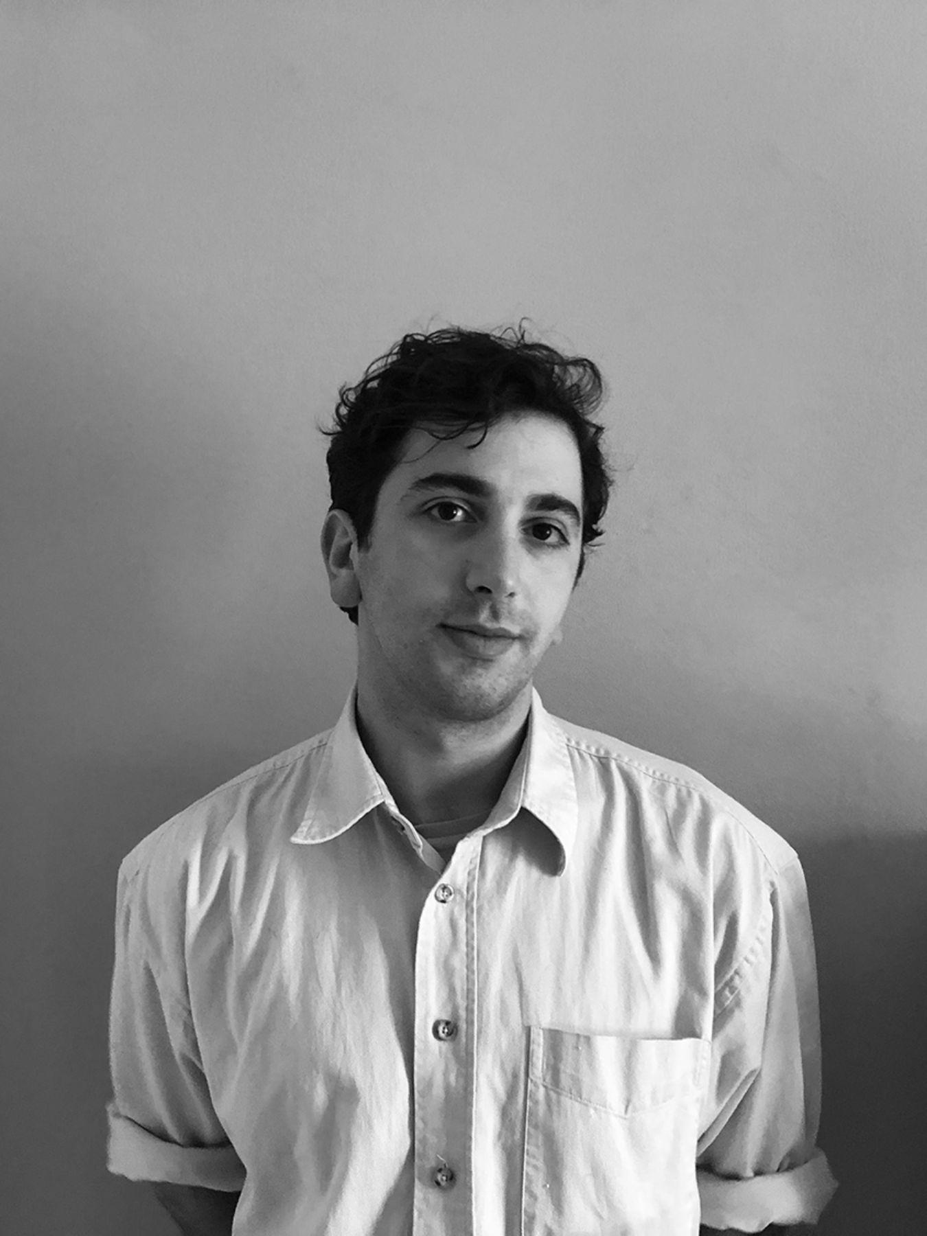 Mario Banushi lächelt sanft. Er trägt ein lockeres Hemd und verschränkt die Hände hinter dem Rücken.