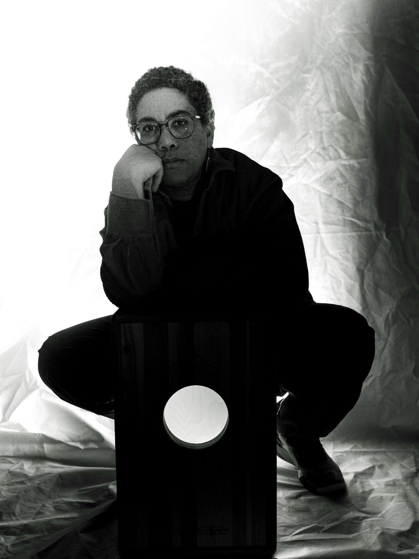 Laura Robles is sitting behind a cajon, her cheek resting on her hand. Robles has short hair and wears glasses.