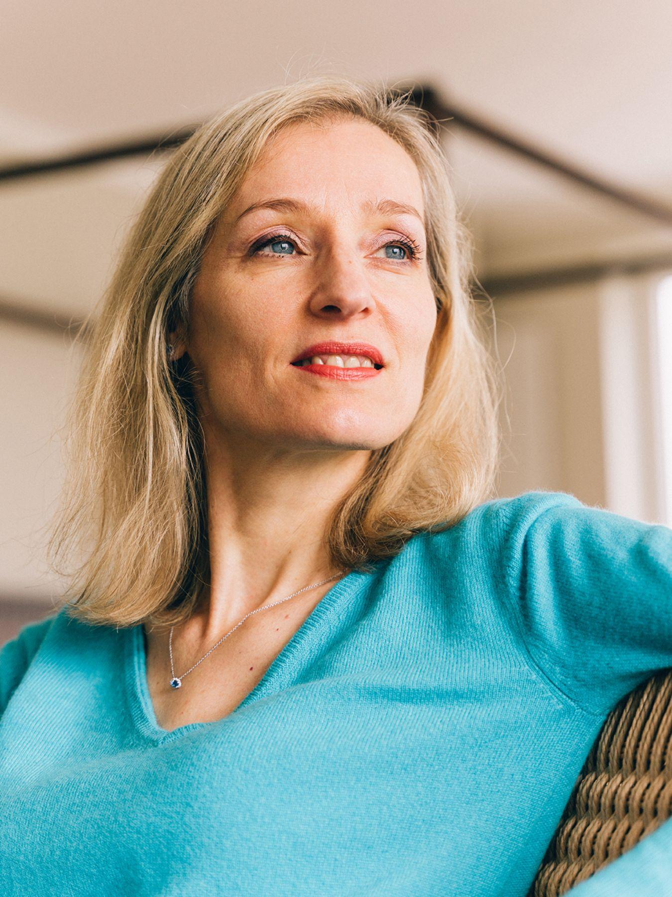 Person mit hellblauem Oberteil und blonden halblangen Haaren in einem Raum.