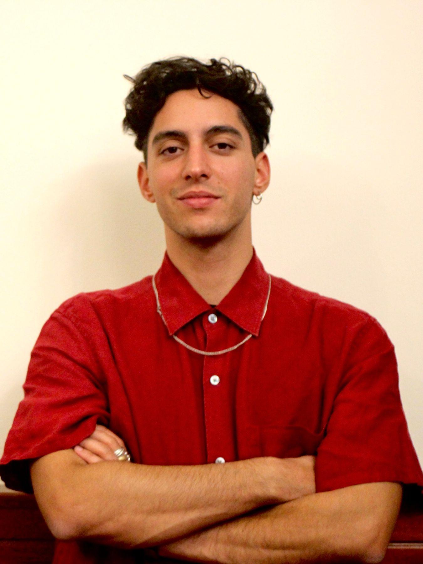 A man in a red shirt with his arms crossed in front of a neutral background