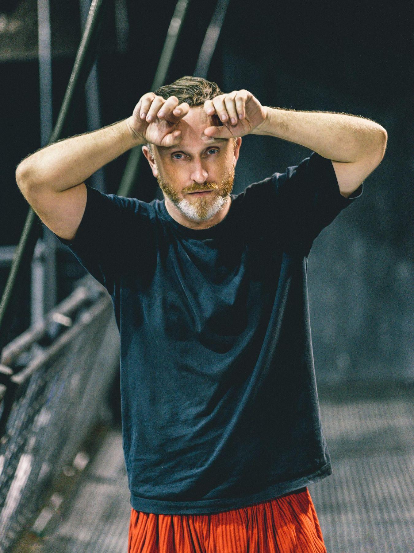 Damien Jalet touches his forehead with the backs of both hands. He is wearing casual clothes and has a full beard.