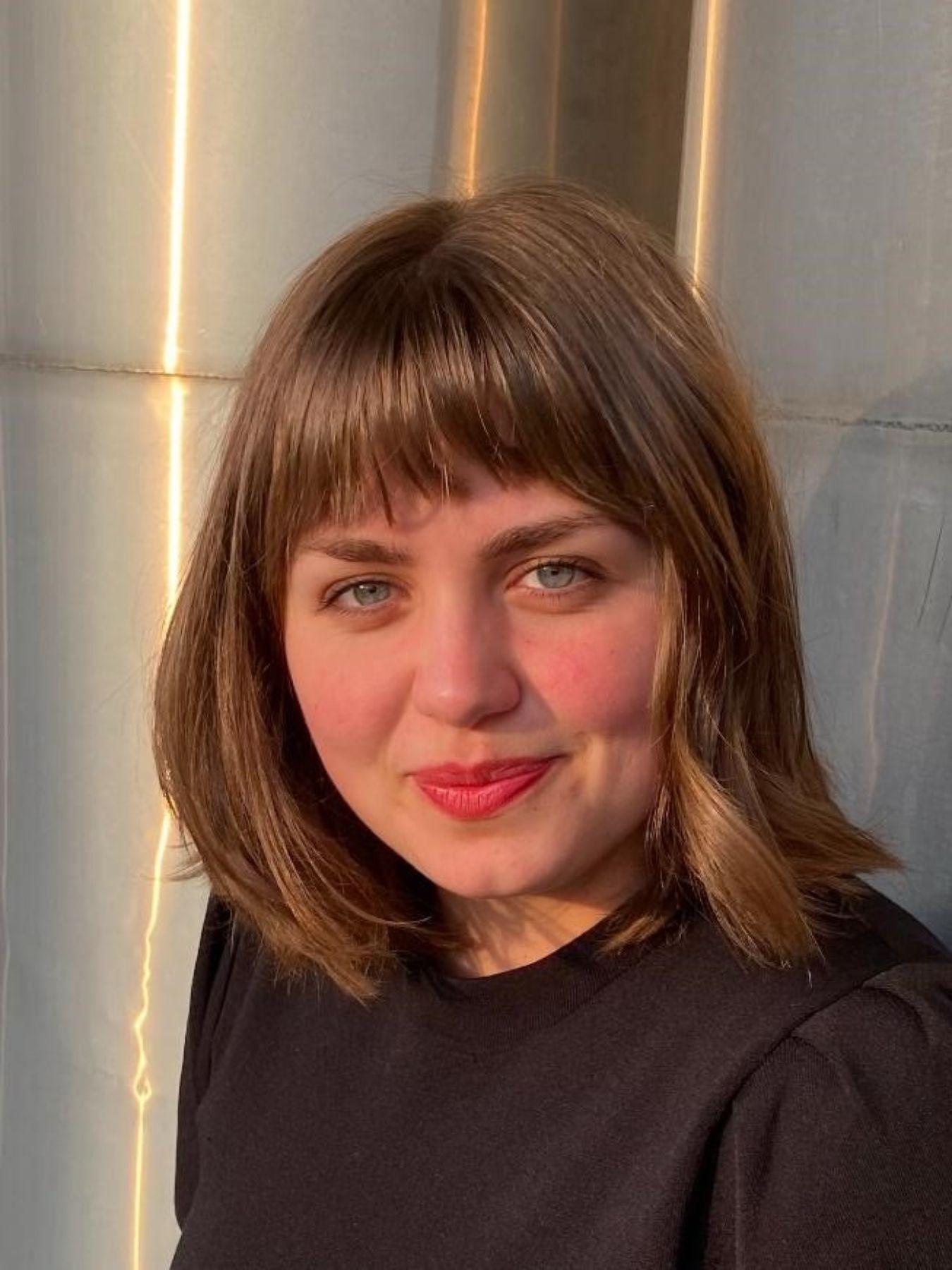 Eine Person mit mittellangen blonden Haaren, schwarzer Bluse und rotem Lippenstift.