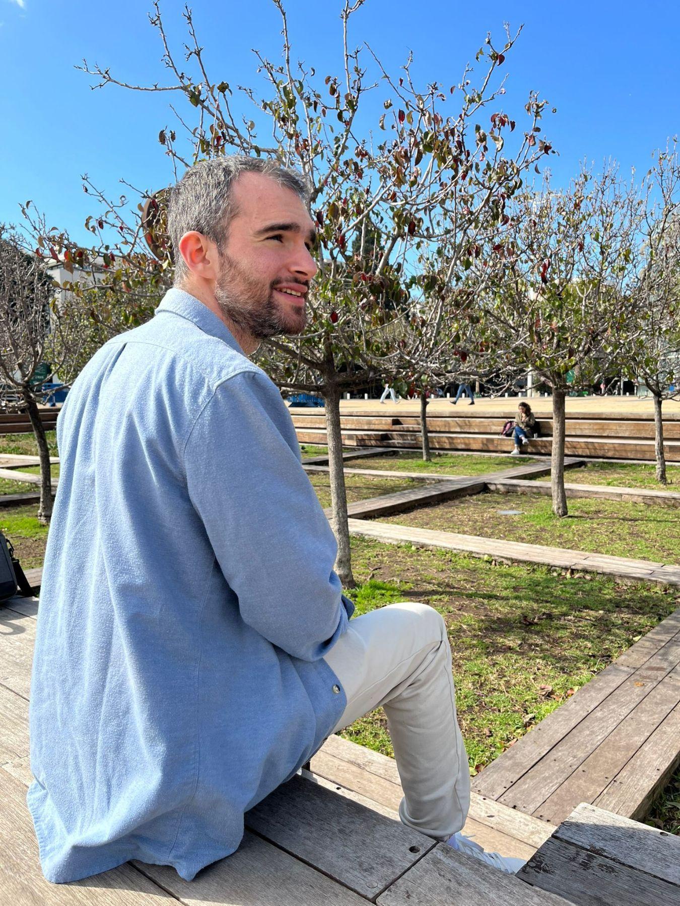 A man in a denim shirt with grey hair and a beard sitting backwards on a wooden platform in front of a tree and looking to the right