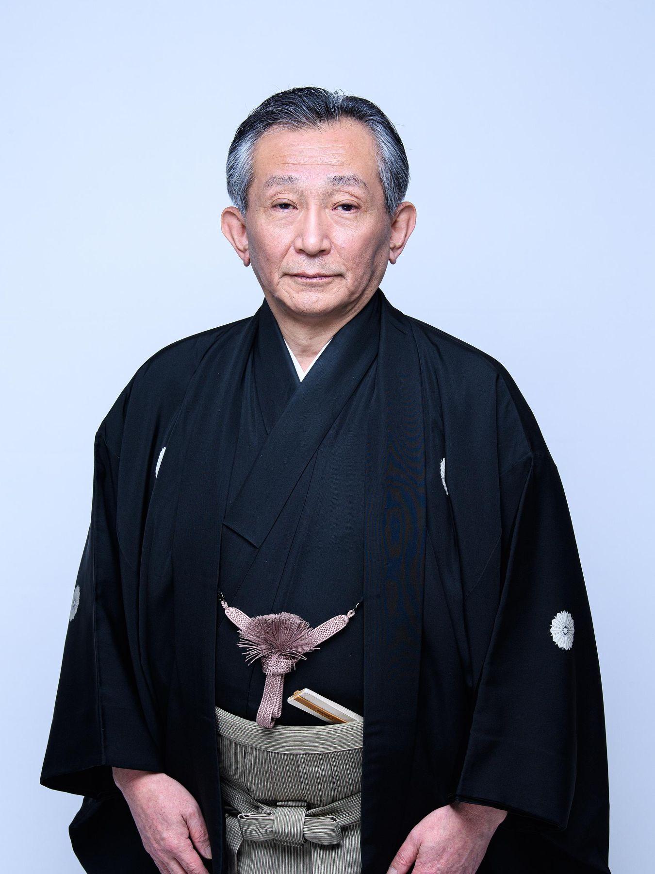 Porträt einer Person im traditionellen japanischen Gewand, stehend vor hellem, einfarbigem Hintergrund, mit dunklem Obergewand und gemustertem Gürtel.