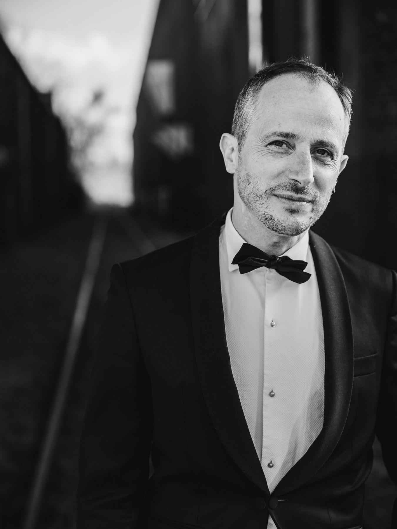 Ein Mann im schwarzen Anzug und mit schwarzer Fliege geht lächend auf die Kamera zu.