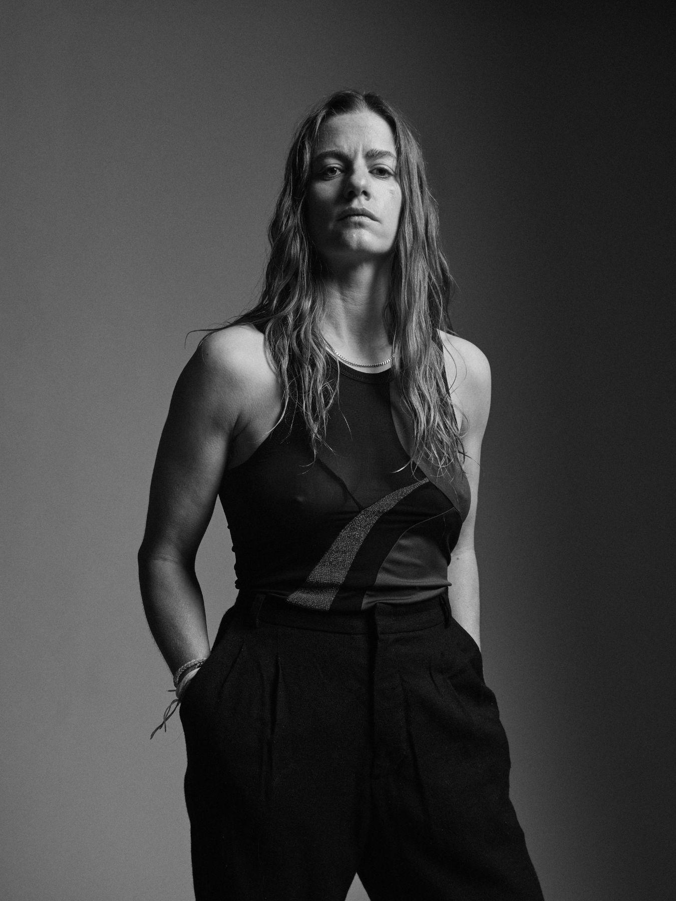 Person in dunklem Outfit steht vor neutralem Hintergrund, Hände in den Hosentaschen.